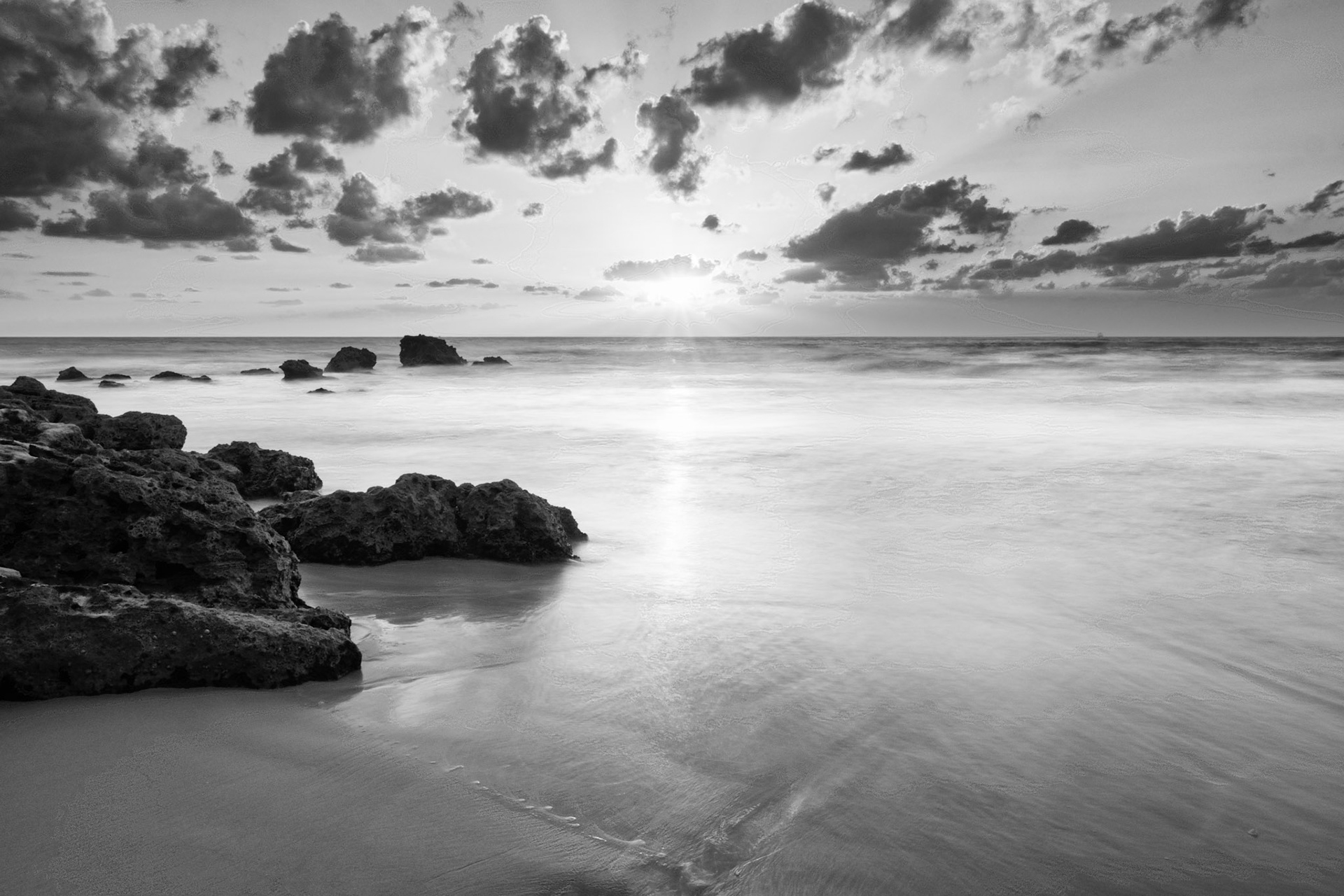 Black and white photo of some rocks and the beach during sunset with clouds