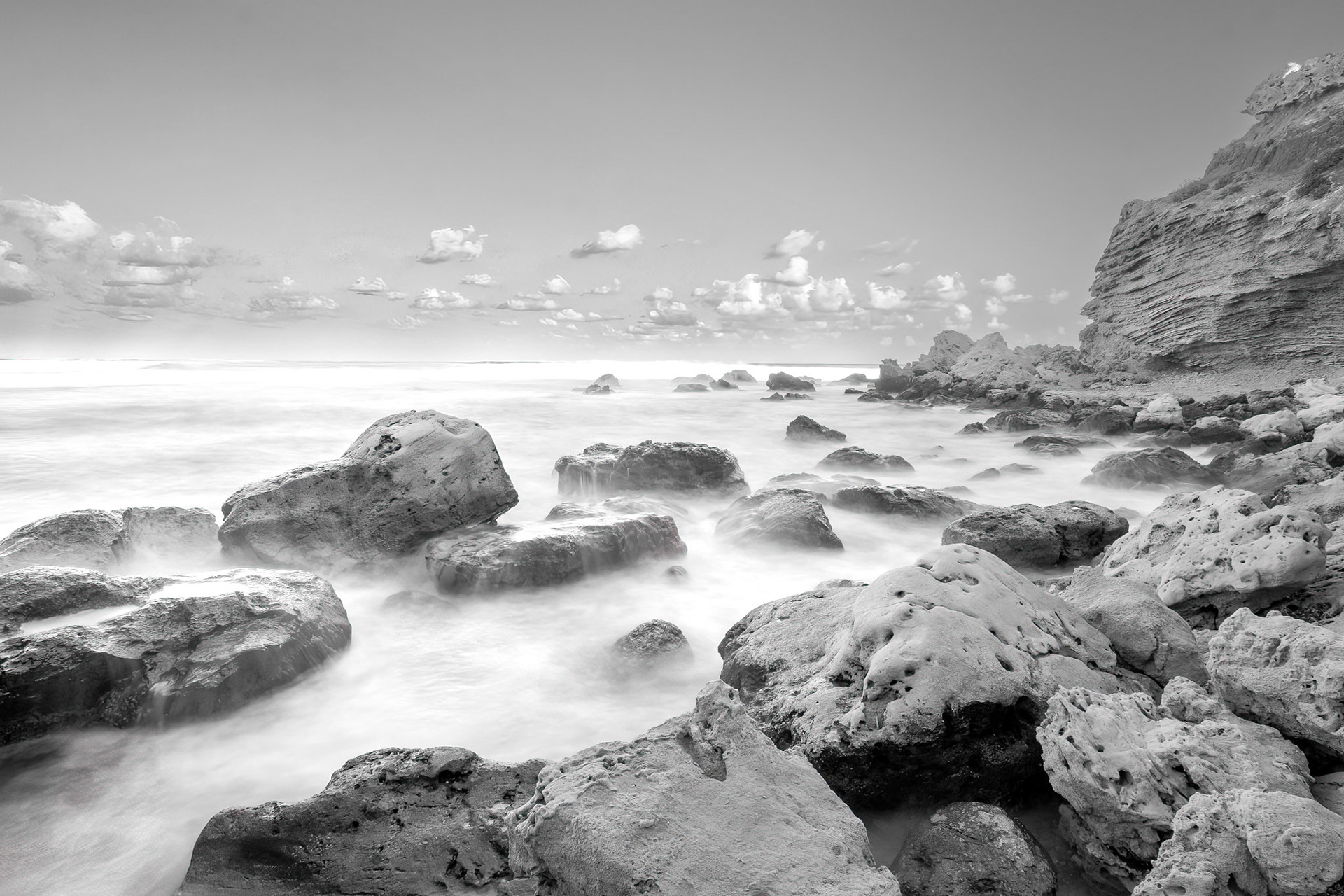 Black and white edit over a long exposure shot of the beach and the rocks formations in it. scattered clouds in the sky.