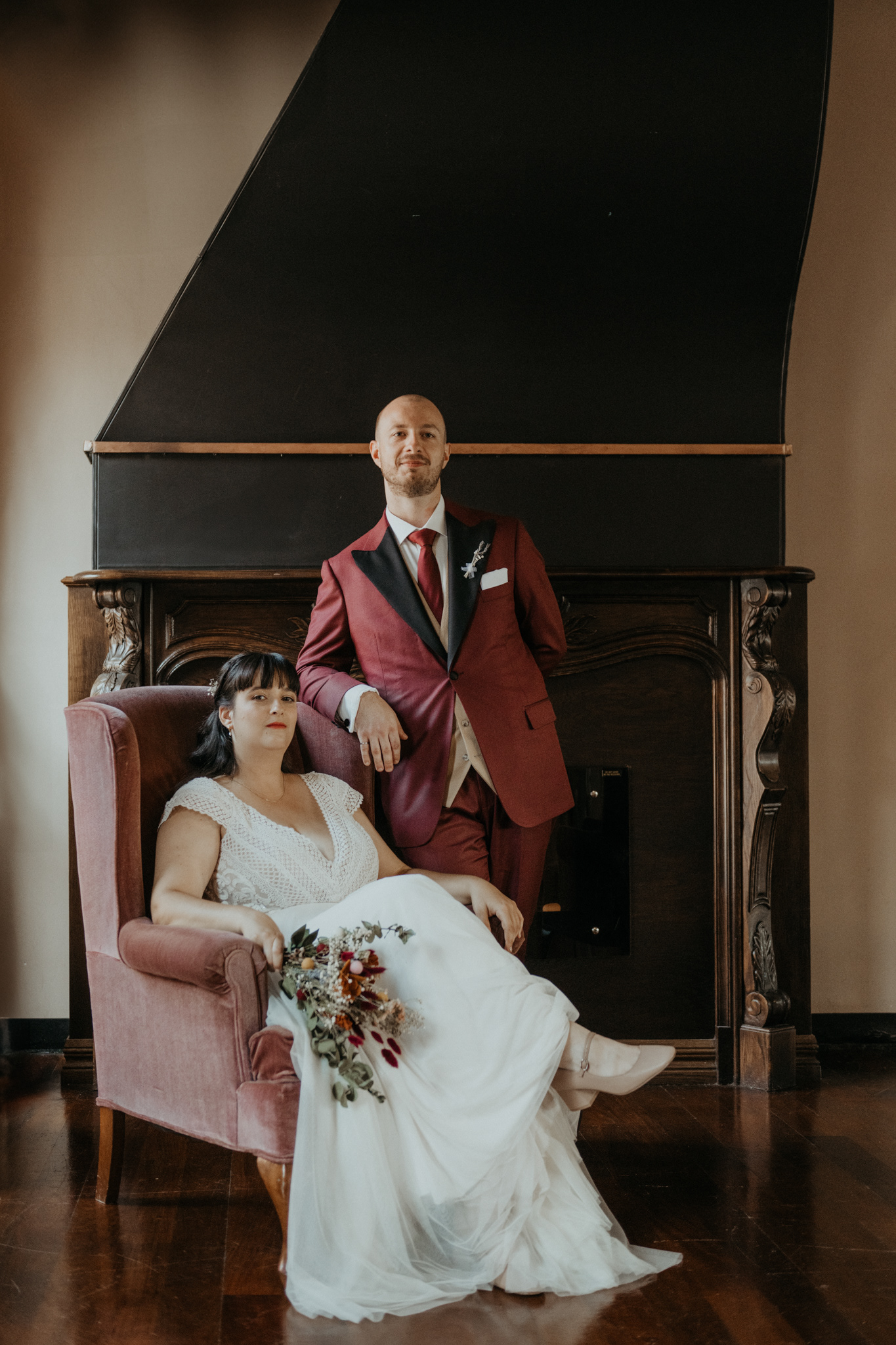 bride sitting on old rose velvet sofa, husband standing, couple looking at the camera, dried flowers, old money, in front of wood fireplace