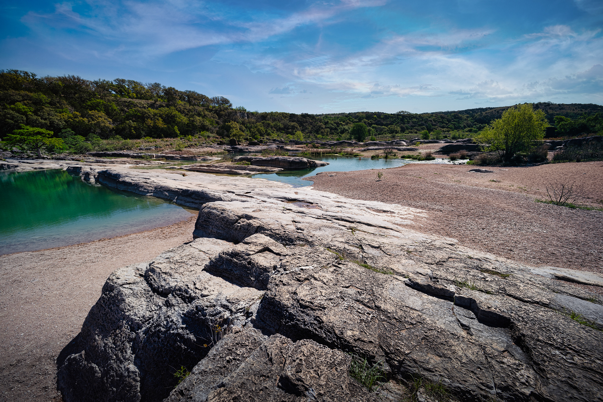 Expansive, sunlit view of Pedernales Falls State Park's Upper Falls in Texas, featuring large, sloping rock slabs leading to pools of water, with tree-lined banks under a blue sky.