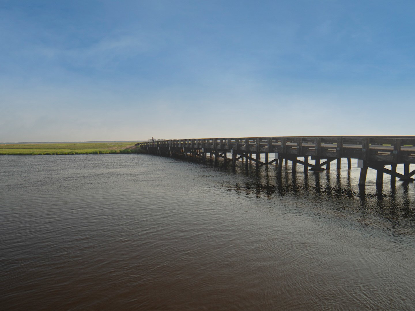 A picturesque scene in Lewes, Delaware, likely featuring a small bridge, possibly wooden, spanning a waterway or marshy area, with coastal vegetation and the town's charming atmosphere suggested.