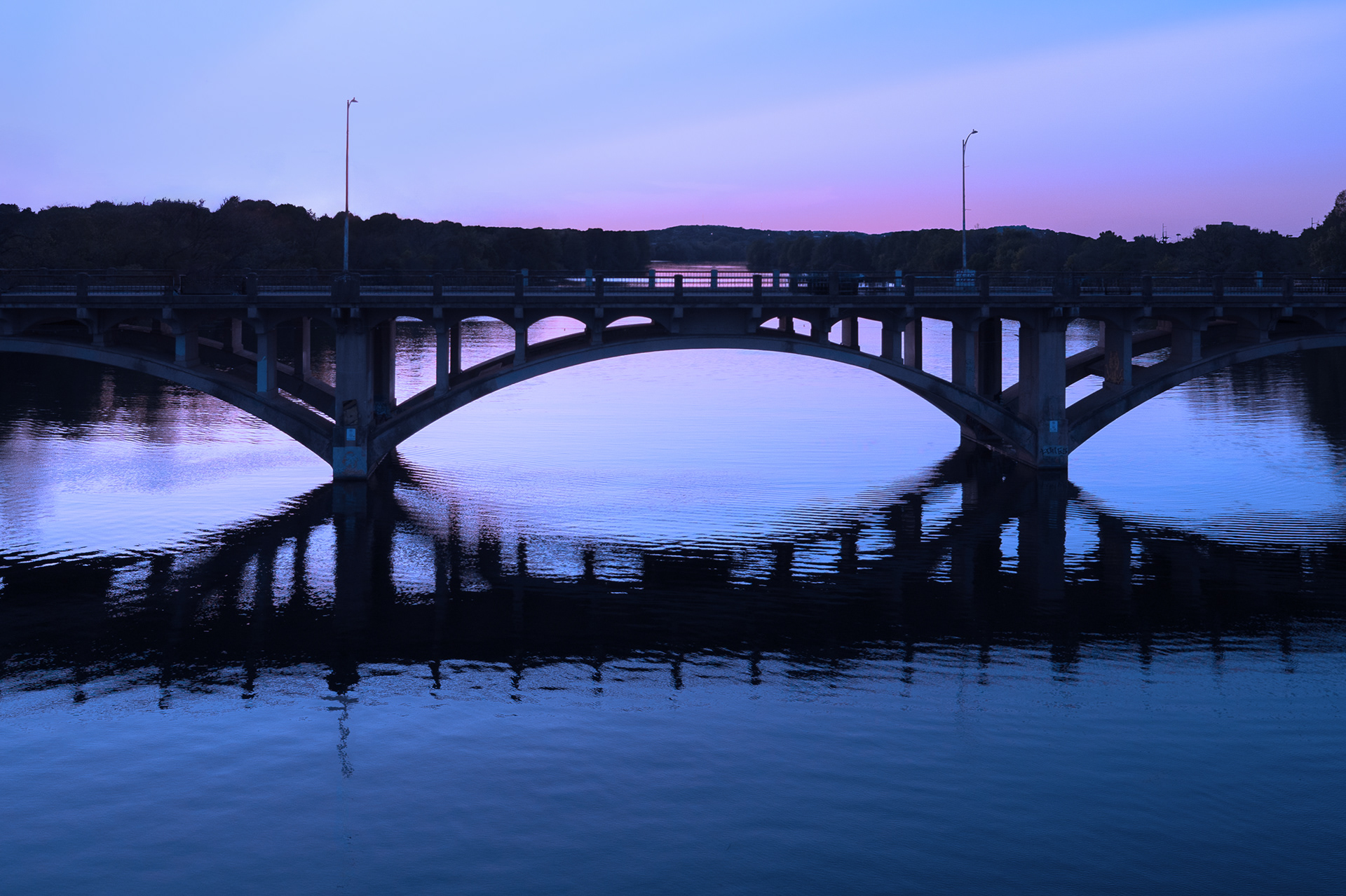 The Lamar Boulevard Bridge in Austin, Texas, an arched concrete structure, reflecting in the calm water below under a purple-hued twilight sky.