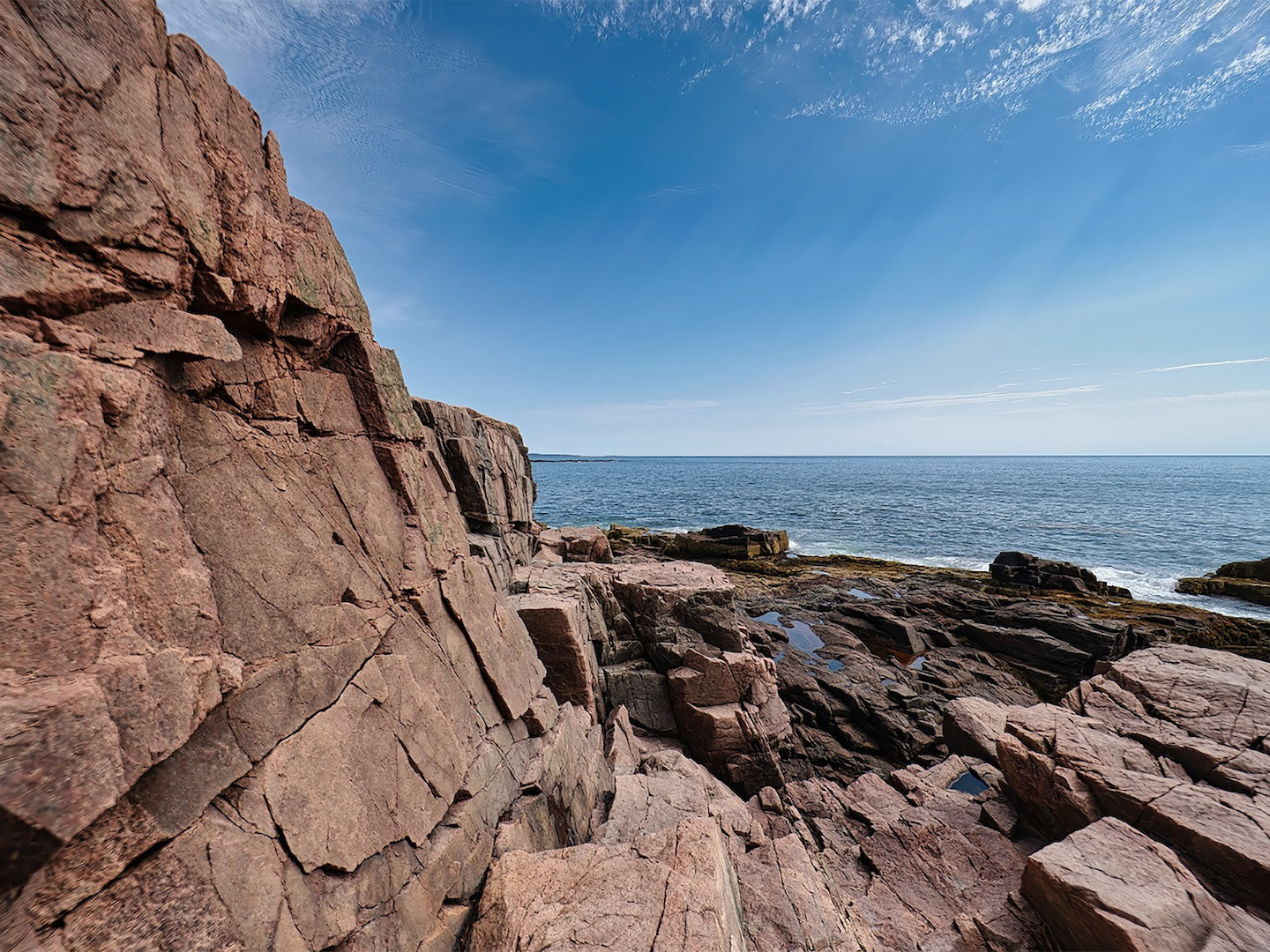 A view of the rugged, rocky shoreline of Acadia National Park, Maine, showcasing granite formations meeting the ocean, possibly with waves crashing and evergreen trees lining the coast.