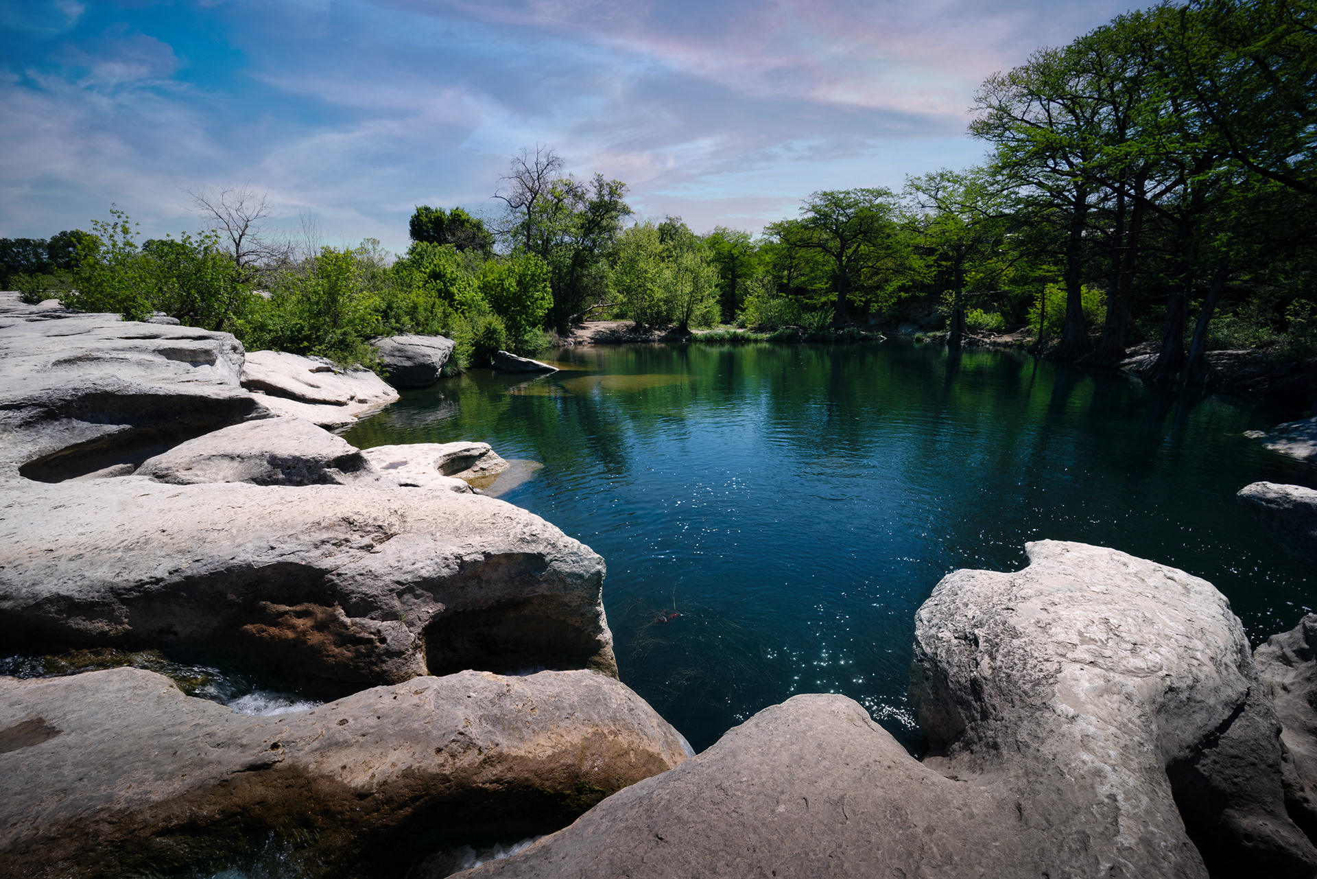 Sun-dappled view of the Upper Falls at McKinney Falls State Park, Texas, showcasing smooth, large rocks bordering a calm, dark pool of water, with lush green trees in the background under a blue sky.