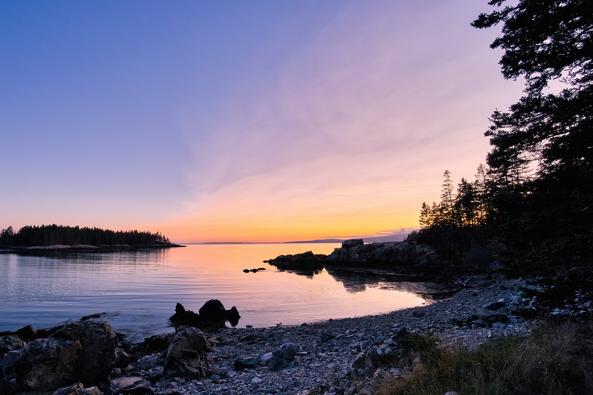 A picturesque sunset over clear water at Schoodic Point in Winter Harbor, Maine, with the scene accented by beautiful trees framing the view.