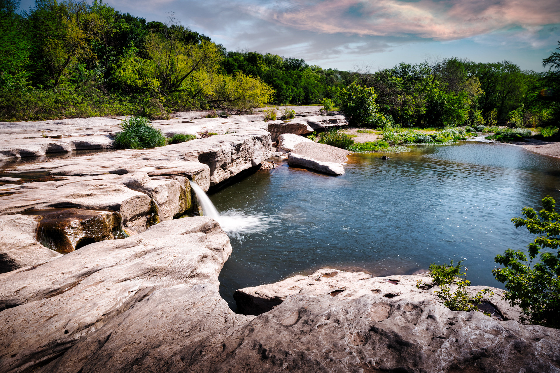 Sunlight illuminates a small waterfall at McKinney Falls State Park, Texas, as water flows over distinctive, smooth rock ledges into a serene pool, surrounded by green trees.