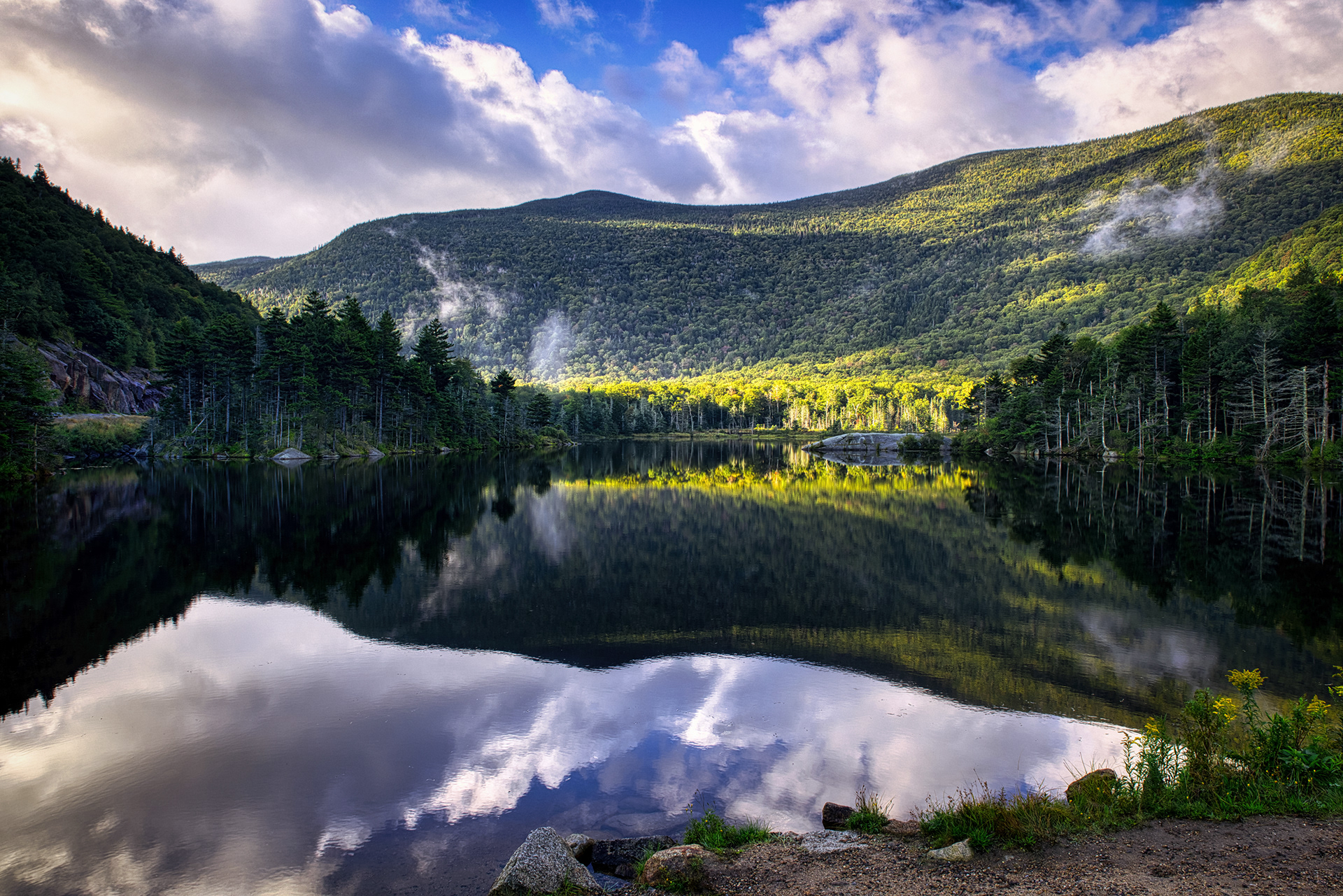 A tranquil scene at Beaver Pond in New Hampshire, likely showing calm waters reflecting surrounding trees, with the White Mountains potentially visible in the background under a clear or partly cloudy sky.