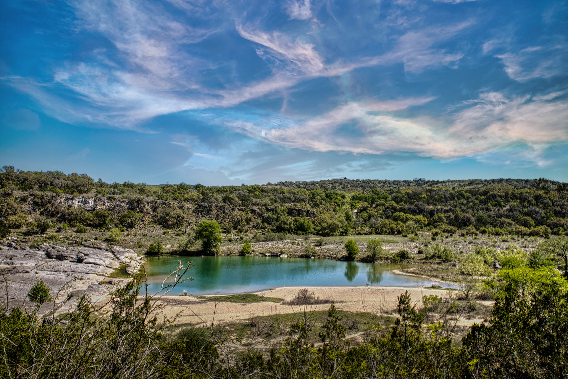 A wide view of the Upper Falls area at Pedernales Falls State Park, Texas, showing a calm blue-green pool of water with a sandy bank, surrounded by rocky terrain and green treed hillsides under a dramatic, colorful sky.