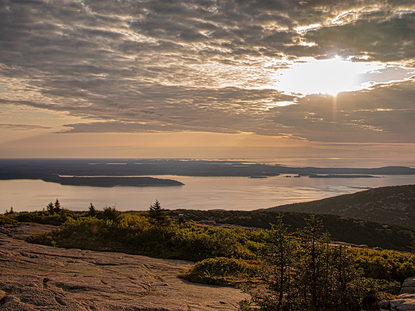 A bright sunset viewed from the summit of Cadillac Mountain in Acadia National Park, Maine, illuminating the landscape with vibrant colors and offering expansive views.