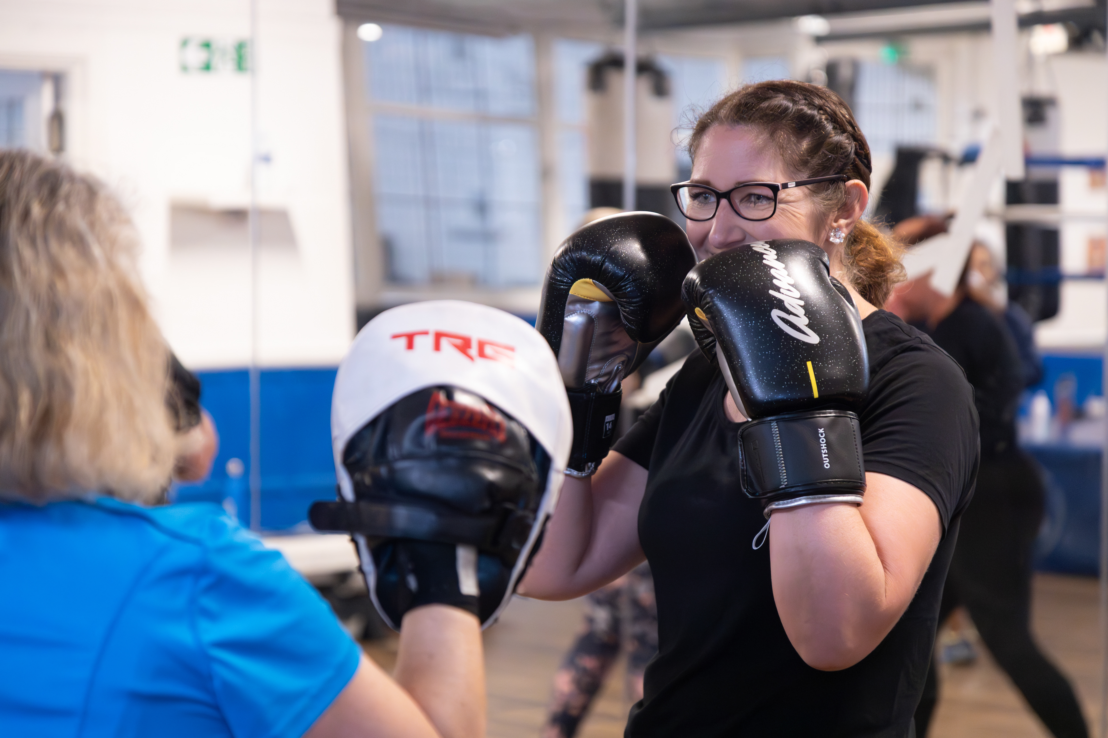 Woman at 12 Rounds boxing club  holds boxing pads for female boxer in black top wearing glasses. Branding photography by Cj Swaby Photography