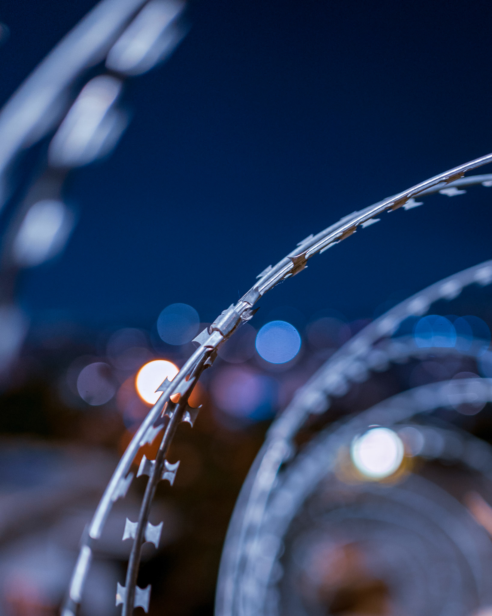 In the city's fuss, there is a strange beauty even in the barriers. Using a shallow #DepthOfField, I wanted to isolate this single twist of #barbwire, turning a harsh industrial line into a piece of floating silver. The city lights in the background dissolve into soft #Bokeh orbs—cyan and amber—creating a dreamlike curtain behind the metal. It’s a reminder that perspective changes everything. What looks like a barrier from afar, becomes a sculpture when you get close enough. A piece for those who appreciate the raw, unpolished side of urban life.🖼️ Industrial elegance for your wall.
