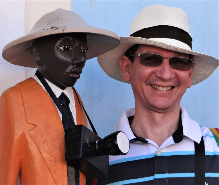 Marcos Cavalcanti usando chapéu claro, óculos escuro e camiseta de gola polo ao lado de uma estátua de um homem negro com uma câmera pendurada ao pescoço