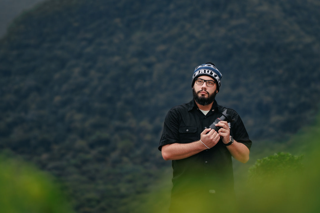 Fotografia de alberth segurando uma câmera com as duas mão, usando touca e uma camisa preta de botões, em um ambiente aparentemente frio com bastante verde