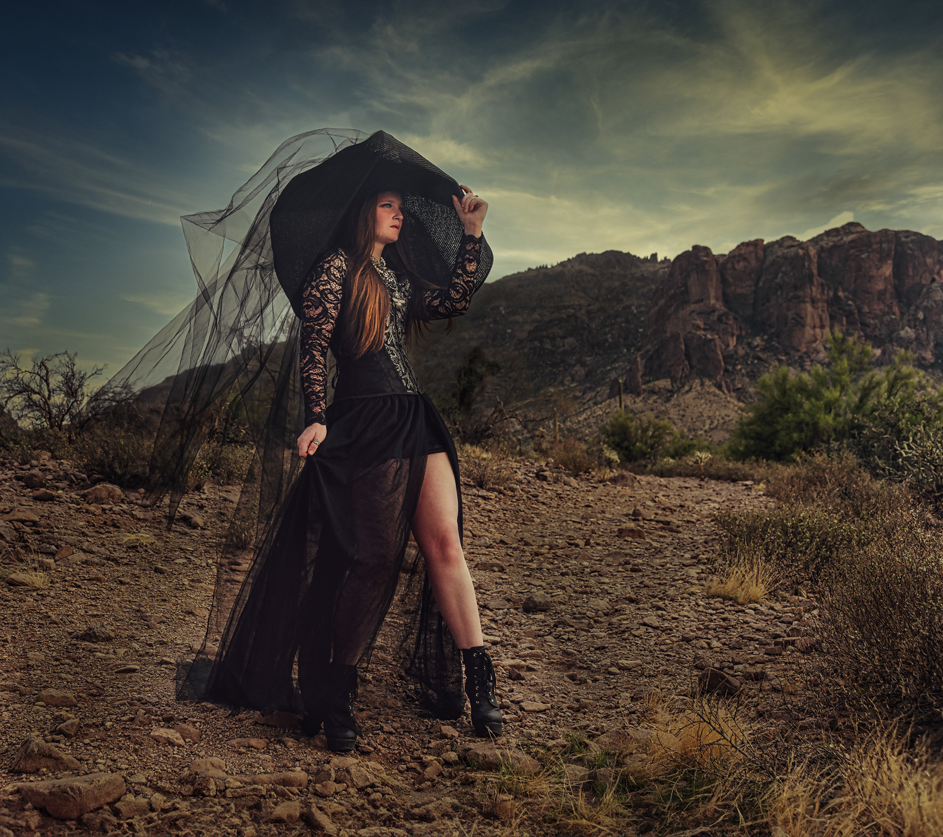 Woman in black tulle dress and wide-brim hat with veil in desert landscape, cinematic fashion portrait