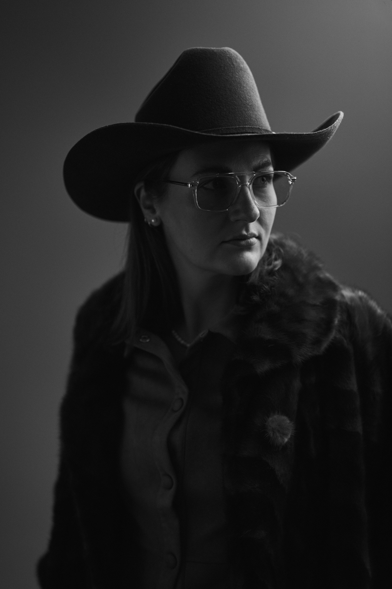 Künstlerisches Schwarz-Weiß-Porträt einer Frau im Studio mit Hut, Brille und Mantel. Minimalistische Portraitfotografie von Michael Kreyer mit weichem Licht und reduzierter Ästhetik.