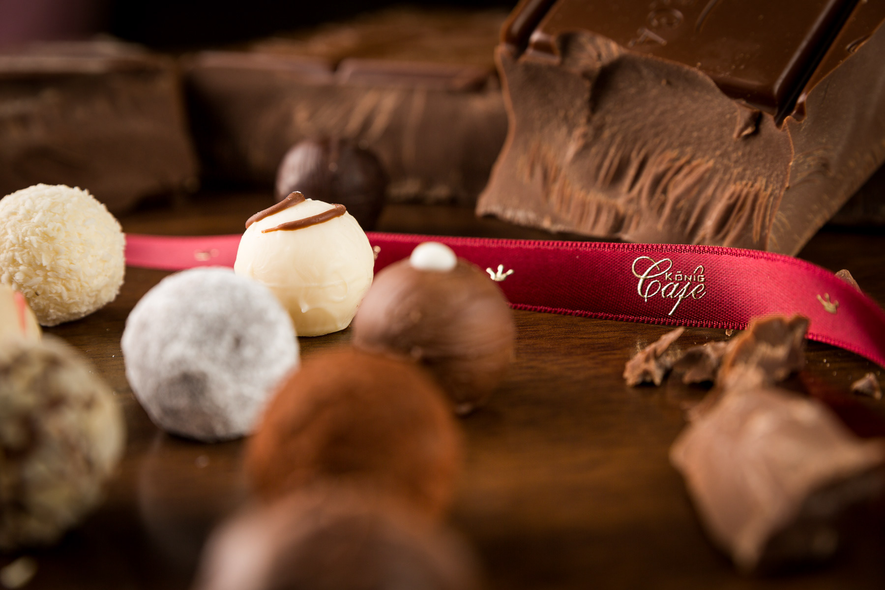 Pralinen im Vordergrund, rotes Band mit Aufdruck verläuft in der Mitte des Bildes, Schokolade im Hintergrund