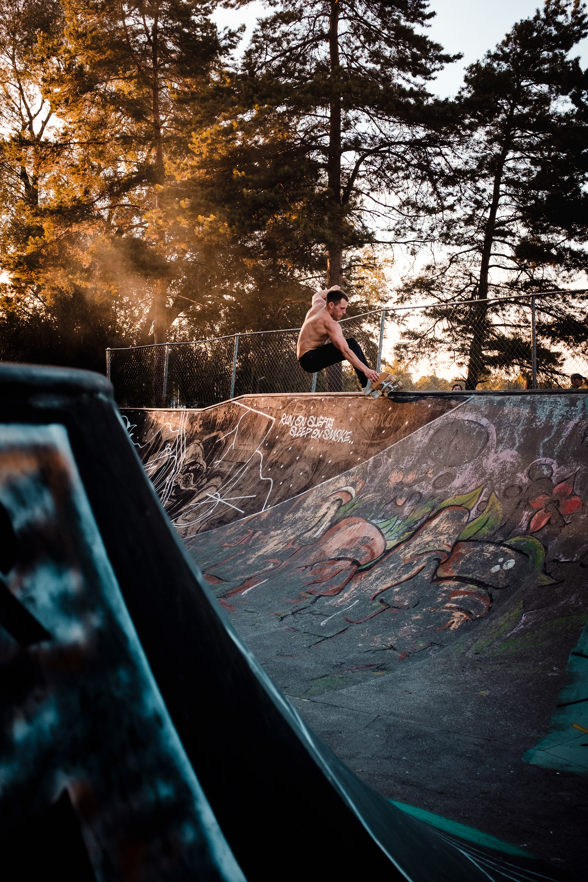 Skateboarder macht Trick mit Grab im Pool, Sonne Rauch und Bäume im Hintergrund