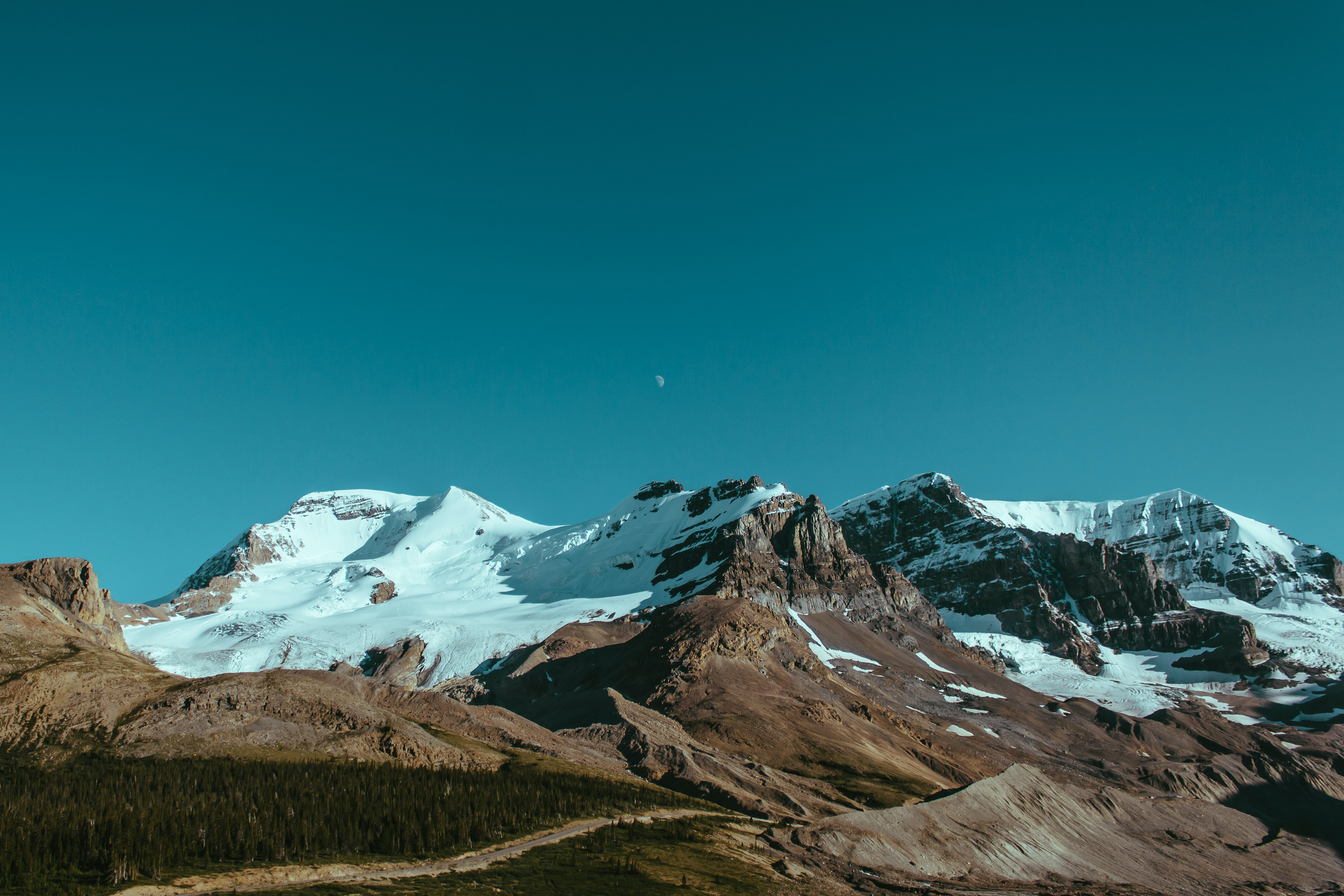 Snow-covered mountain peaks under clear blue sky with faint moon visible.