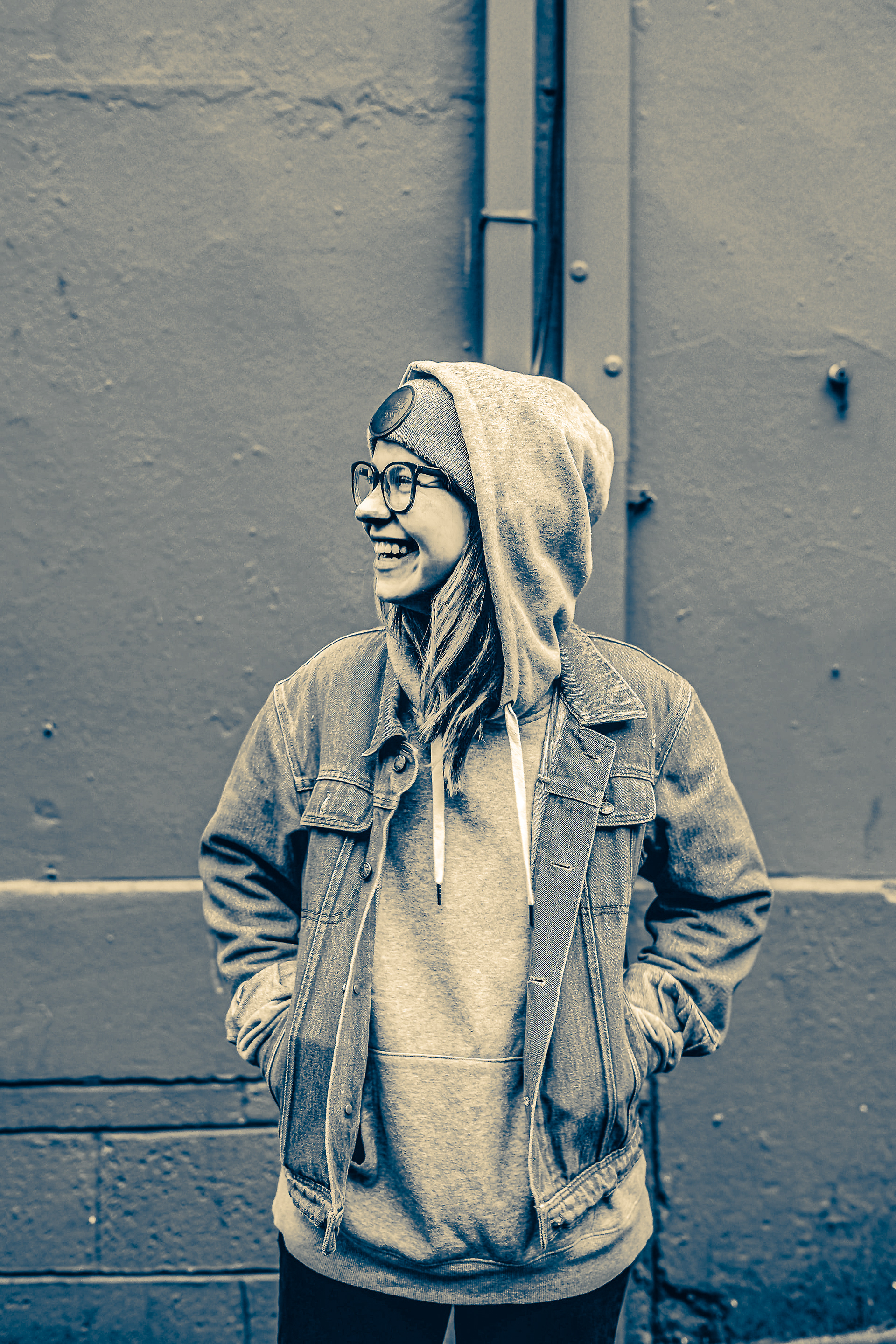 Smiling person in glasses wearing hoodie and denim jacket against wall.