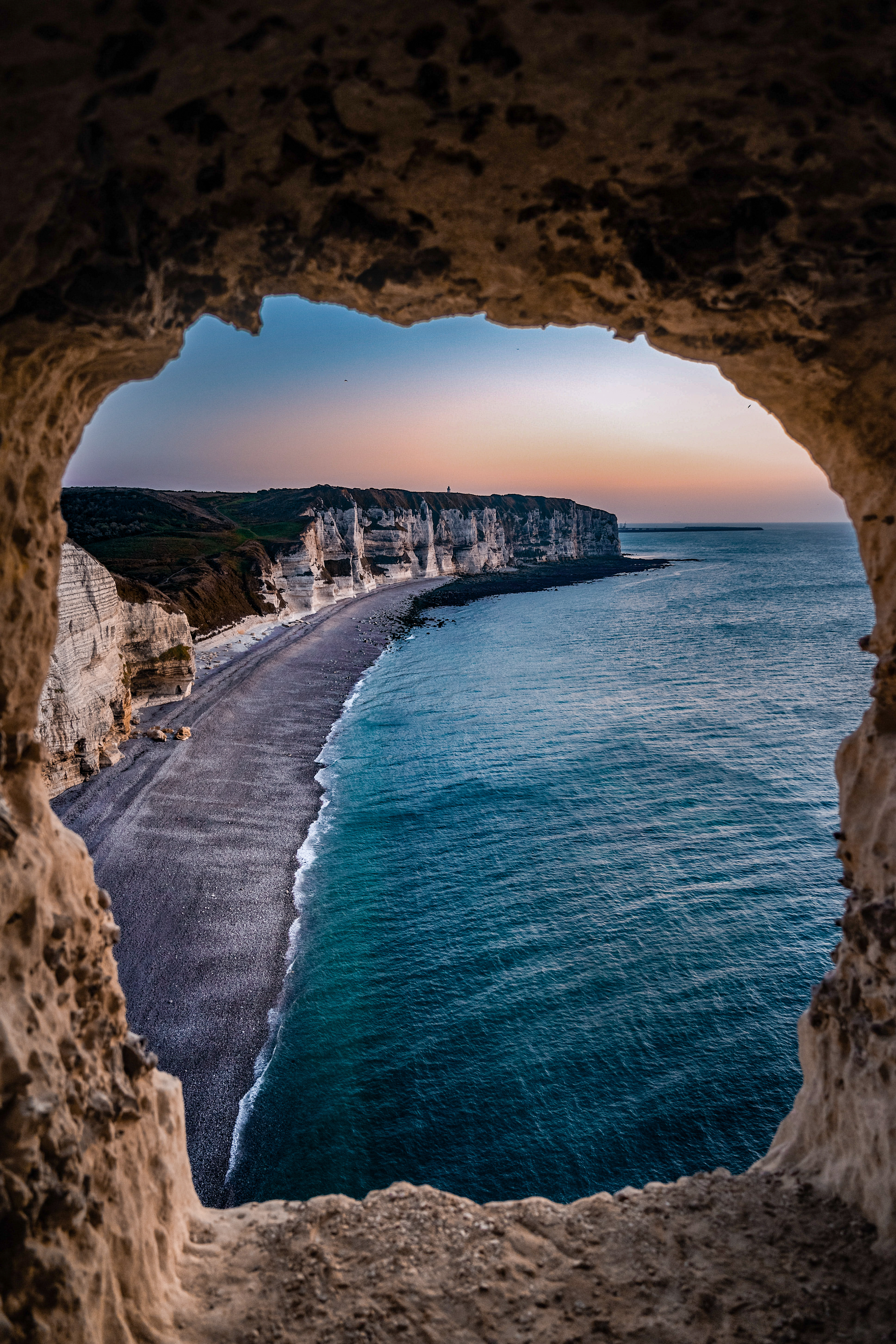 Scenic view of white cliffs and curving shoreline from cave opening with calm sea and pastel sunset sky.