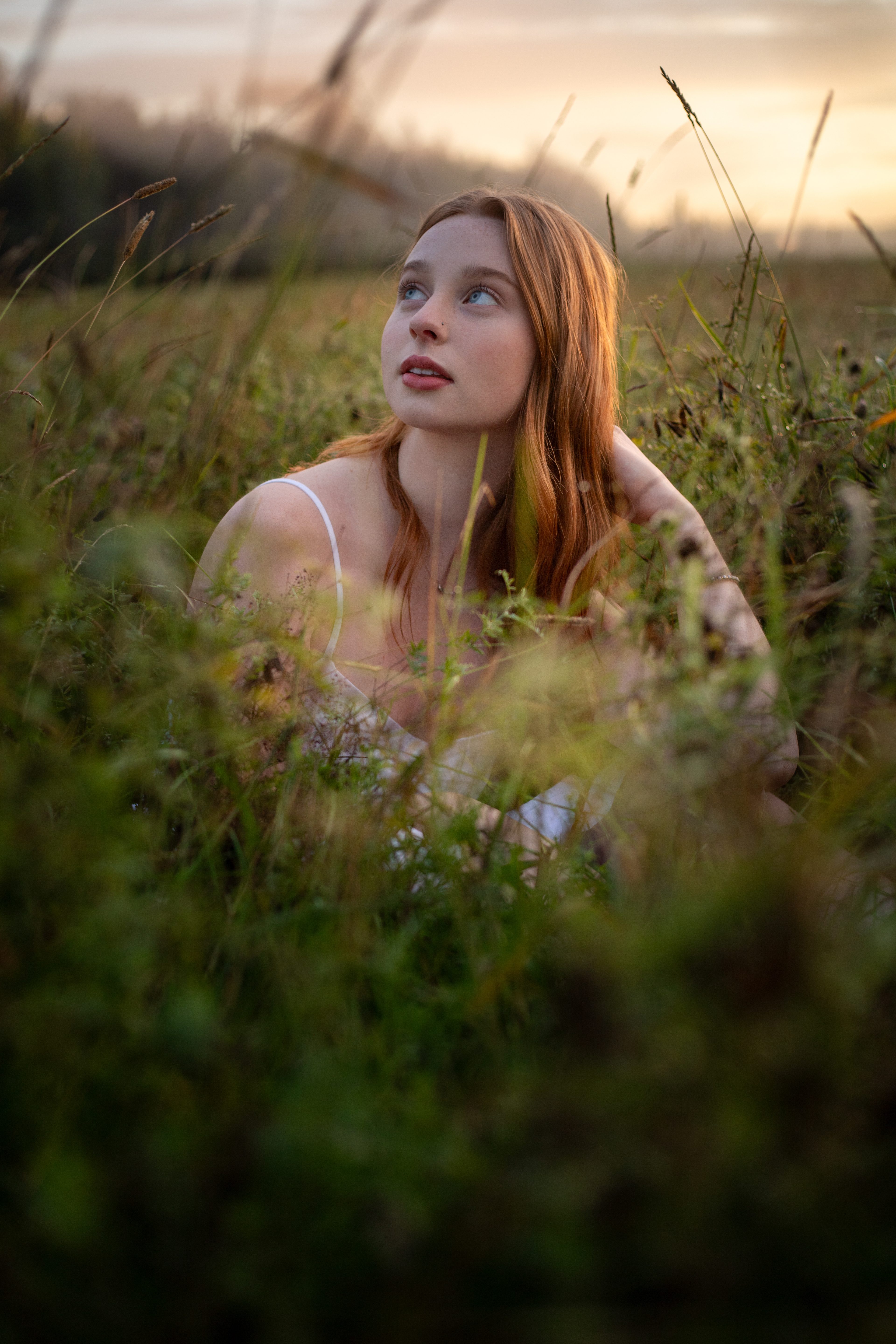 Young woman with long red hair and blue eyes lying in grassy field under golden hour light.