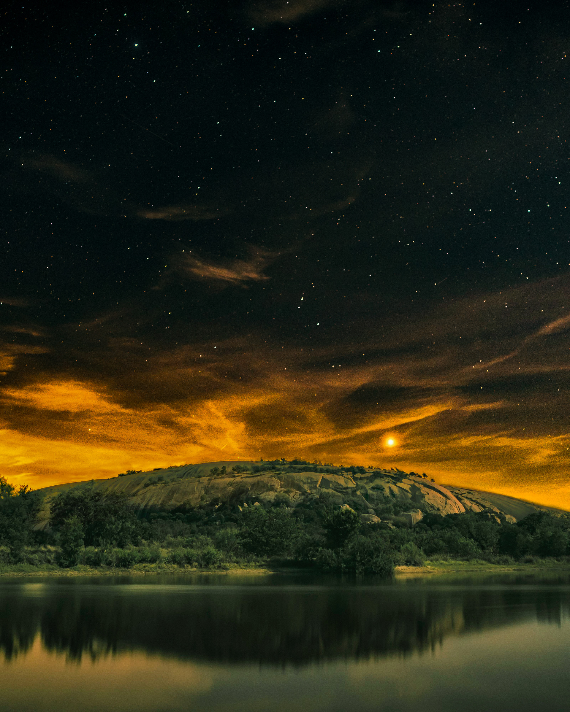 Dramatic landscape with rocky hill, dense trees, and reflective lake under golden night sky with stars and glowing moon.