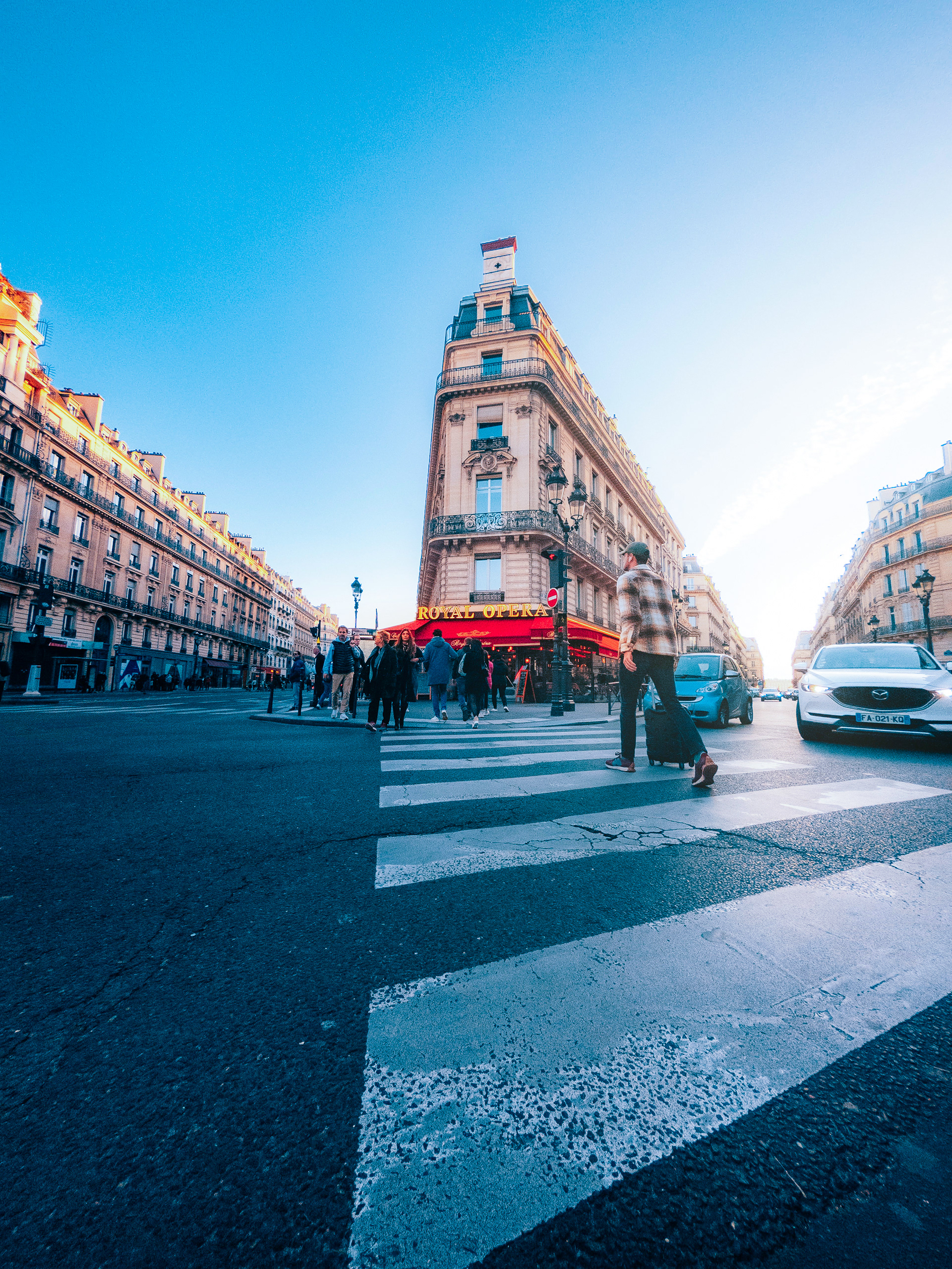 Wide-angle street photo in Paris with pedestrians at zebra crossing, Royal Opera café, cars, and bright blue sky.