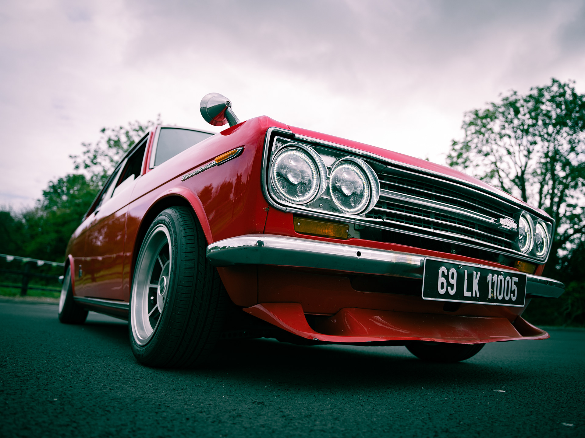 Low-angle photo of red Nissan Bluebird car focusing on front side with headlights and wheels.