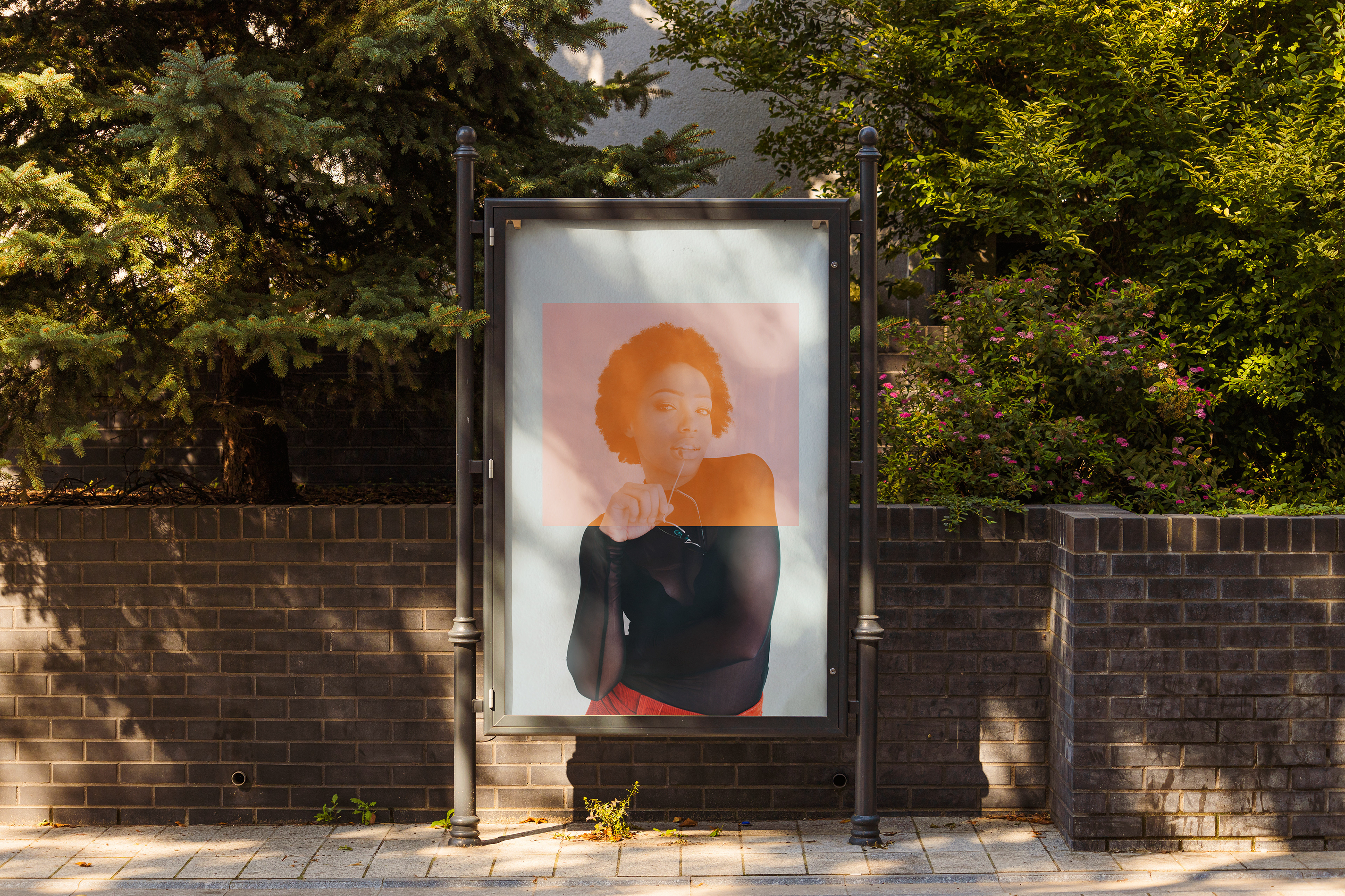 Outdoor poster mockup with portrait of woman and orange overlay in black frame on brick wall with greenery.