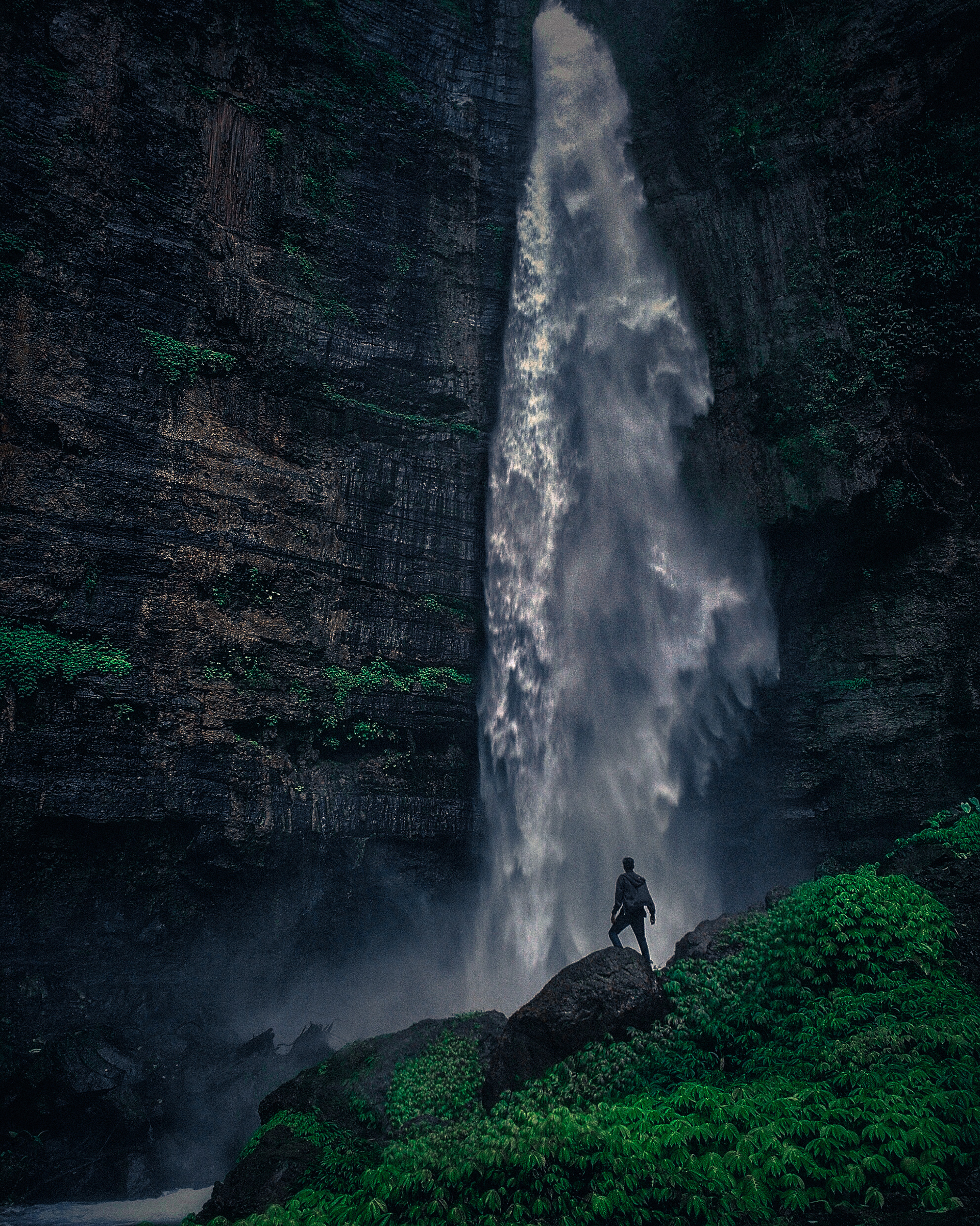 Person standing on rock beneath powerful waterfall cascading down dark cliffs with lush greenery and mist.
