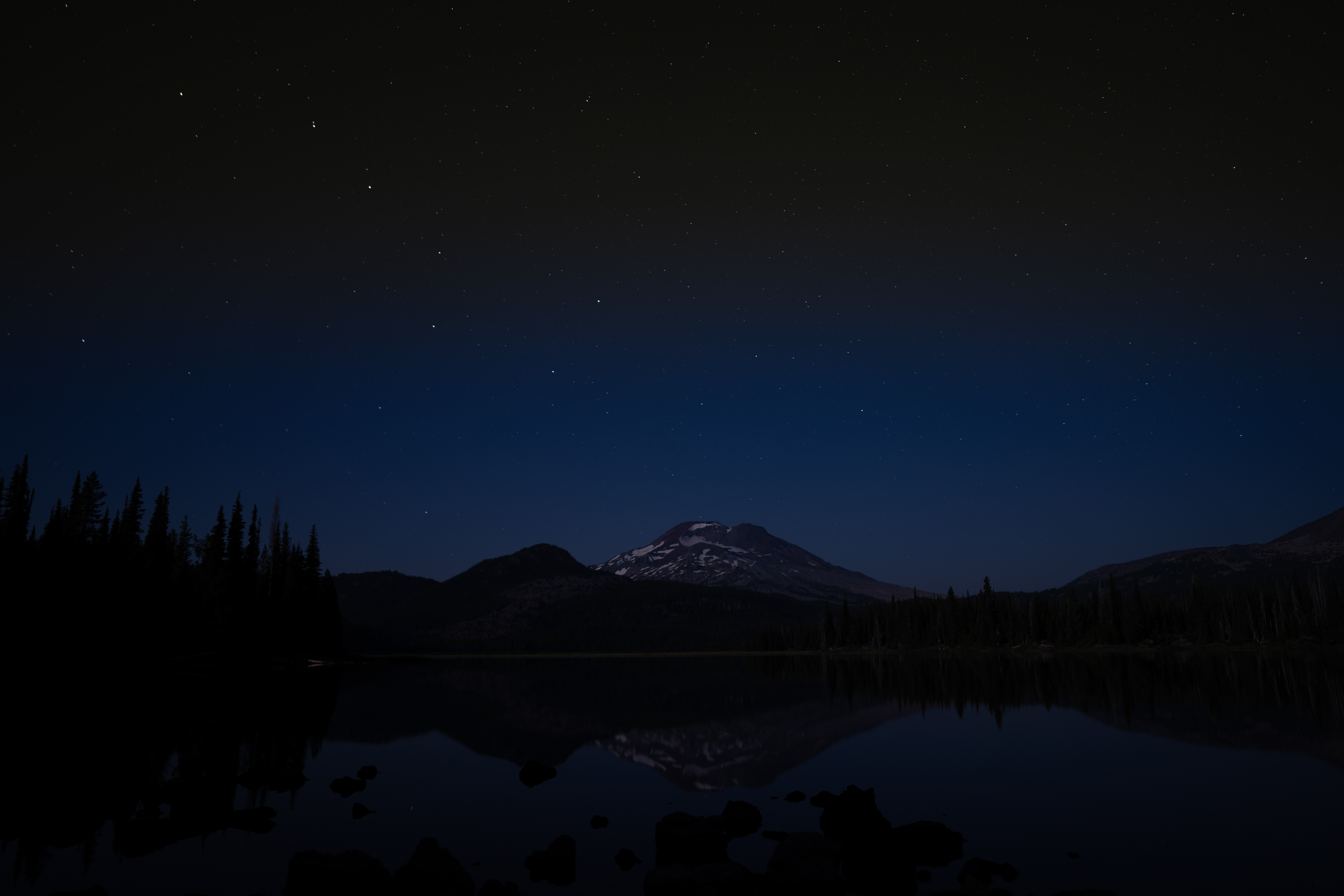 Snowy mountain under starry night sky reflected in still dark lake surrounded by trees.