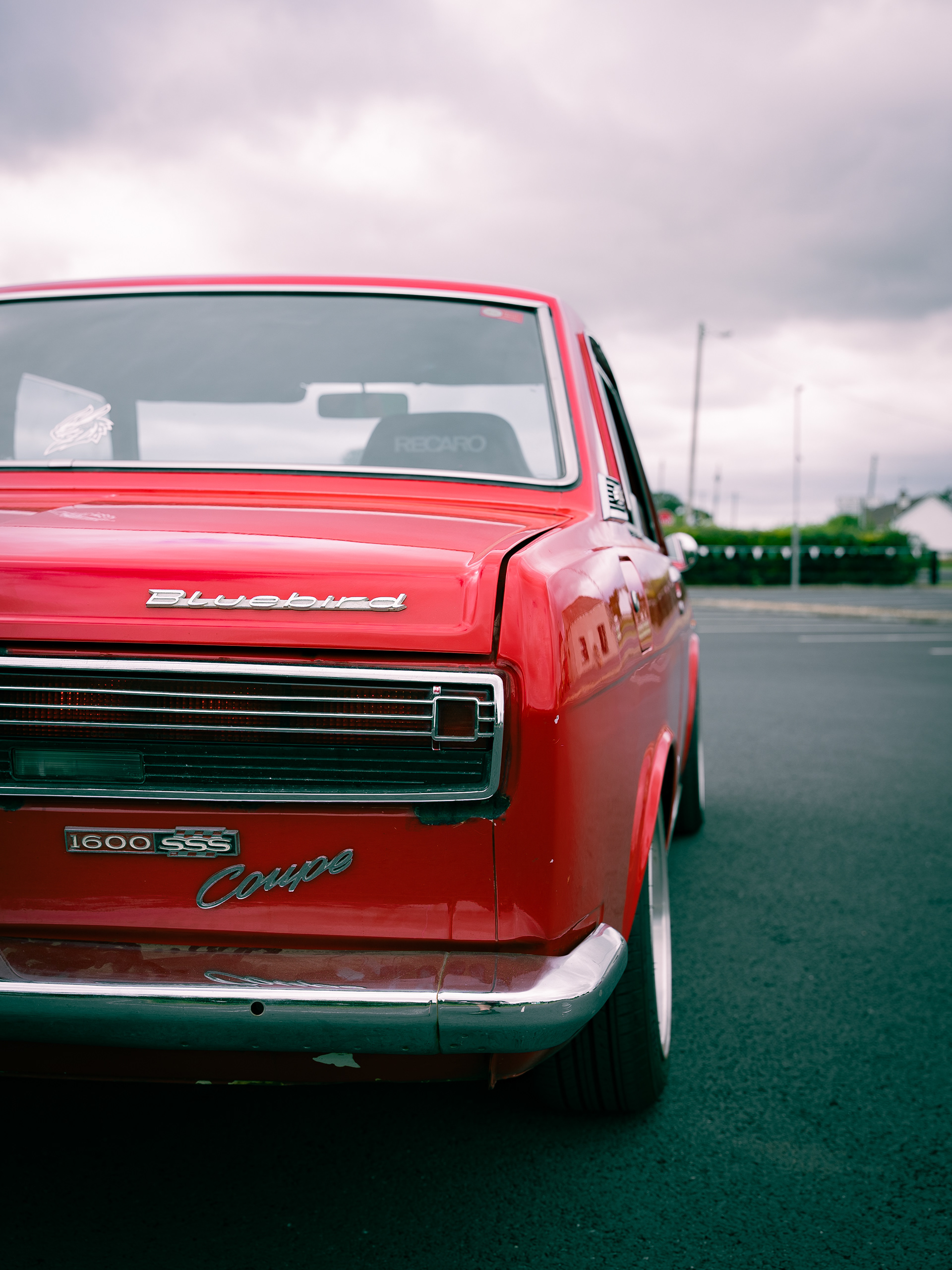 Close-up of silver side mirror mounted on hood of red Nissan Bluebird car.