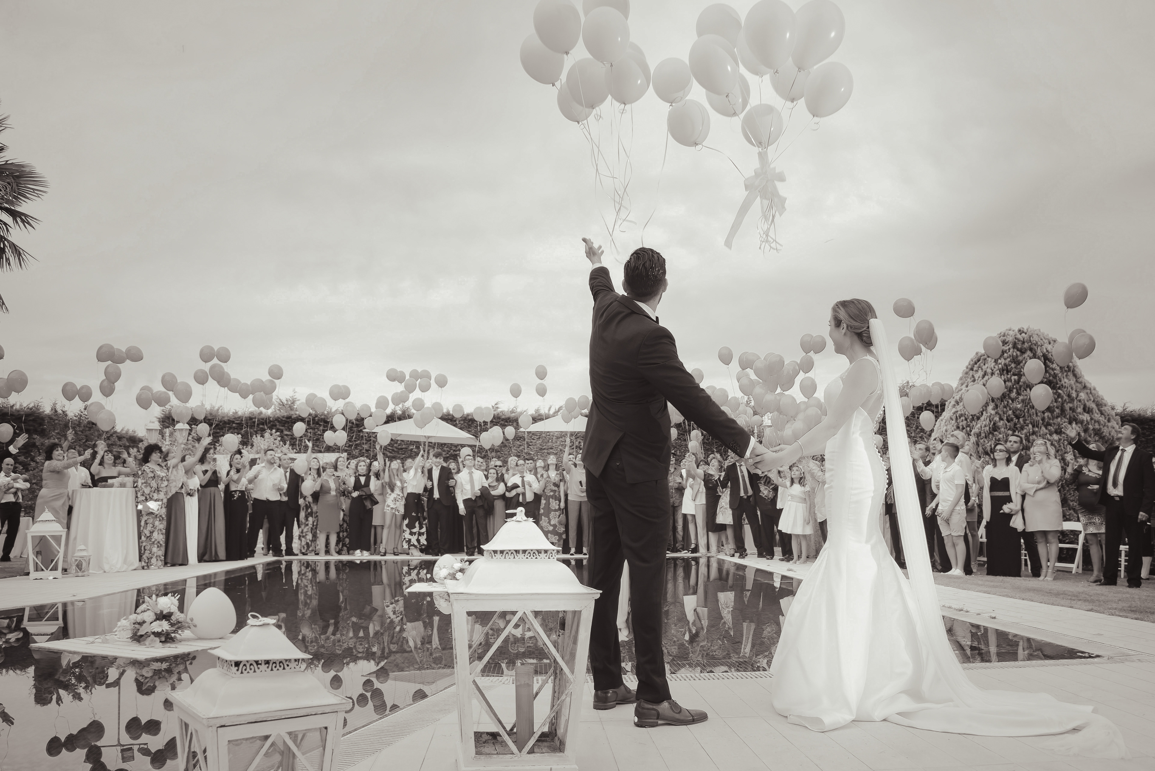 Black-and-white wedding photo of bride and groom releasing balloons with guests outdoors.