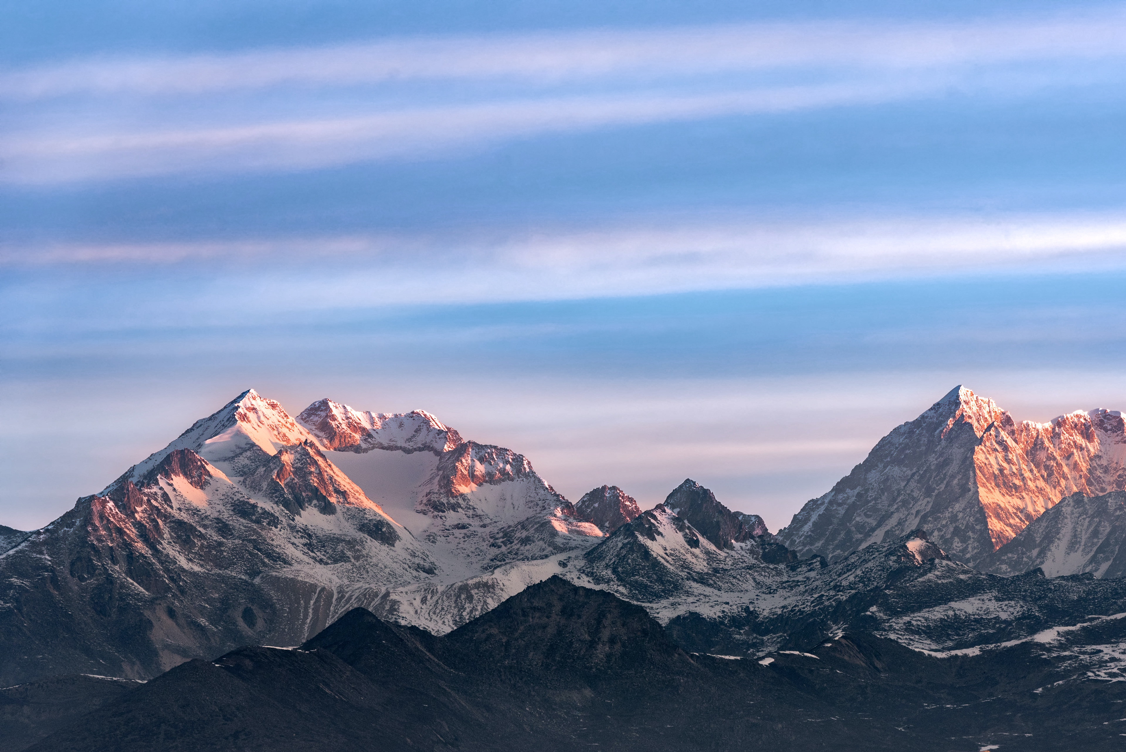 Snow-covered mountain peaks at sunrise with warm glow, clear blue sky, and pink streaks in background.