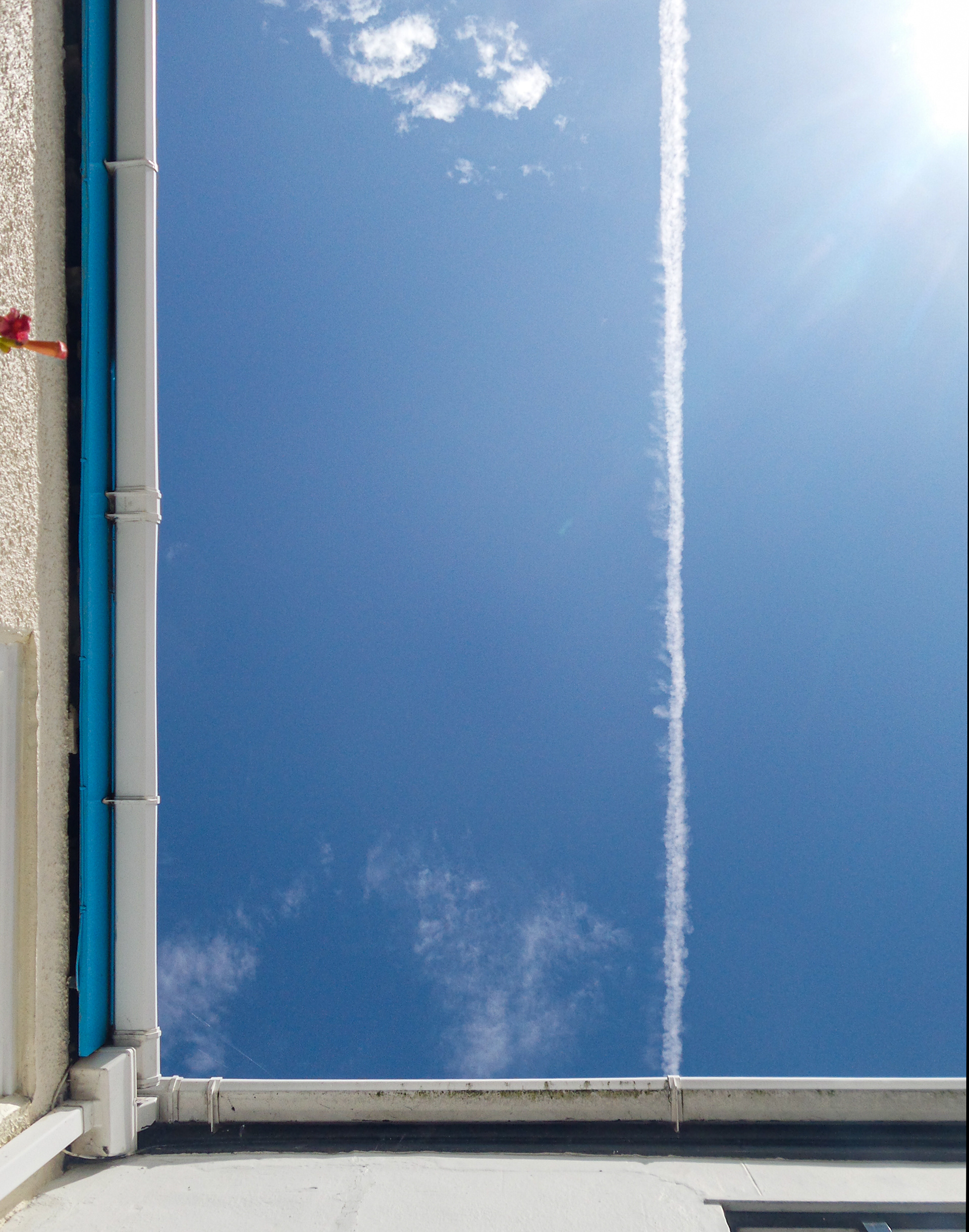 The underside of two white gutters at rightangles are shown against a blue sky with a cloud and vapourtrail. The gutter on the left is clean whilst the one at the bottom has dark marks from algal growth