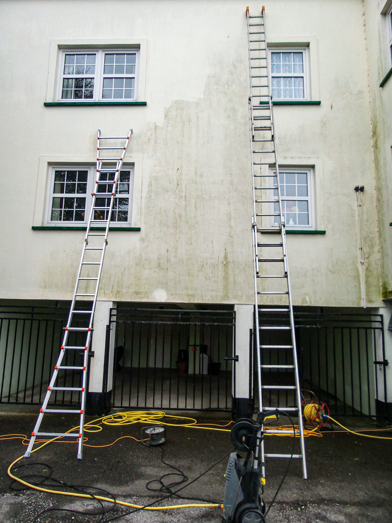 The rendered whiteish wall of a multi-story building is shown with ladders, hoses and powerwasher. To the top left, the wall has been cleaned whilst the rest of the wall is tinged green with algal growth