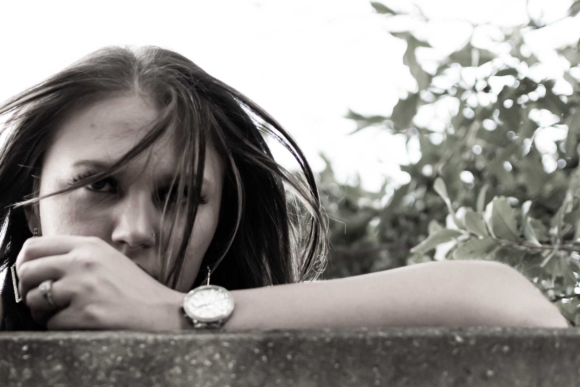 An unsaturated photography of a brunette woman peering thru her hair while wearing a silver watch. The photograph was taken by Tuccera LLC.
