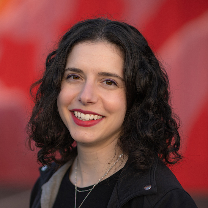 Emily Levine Headshot. Person wearing black smiling in front of red wall.