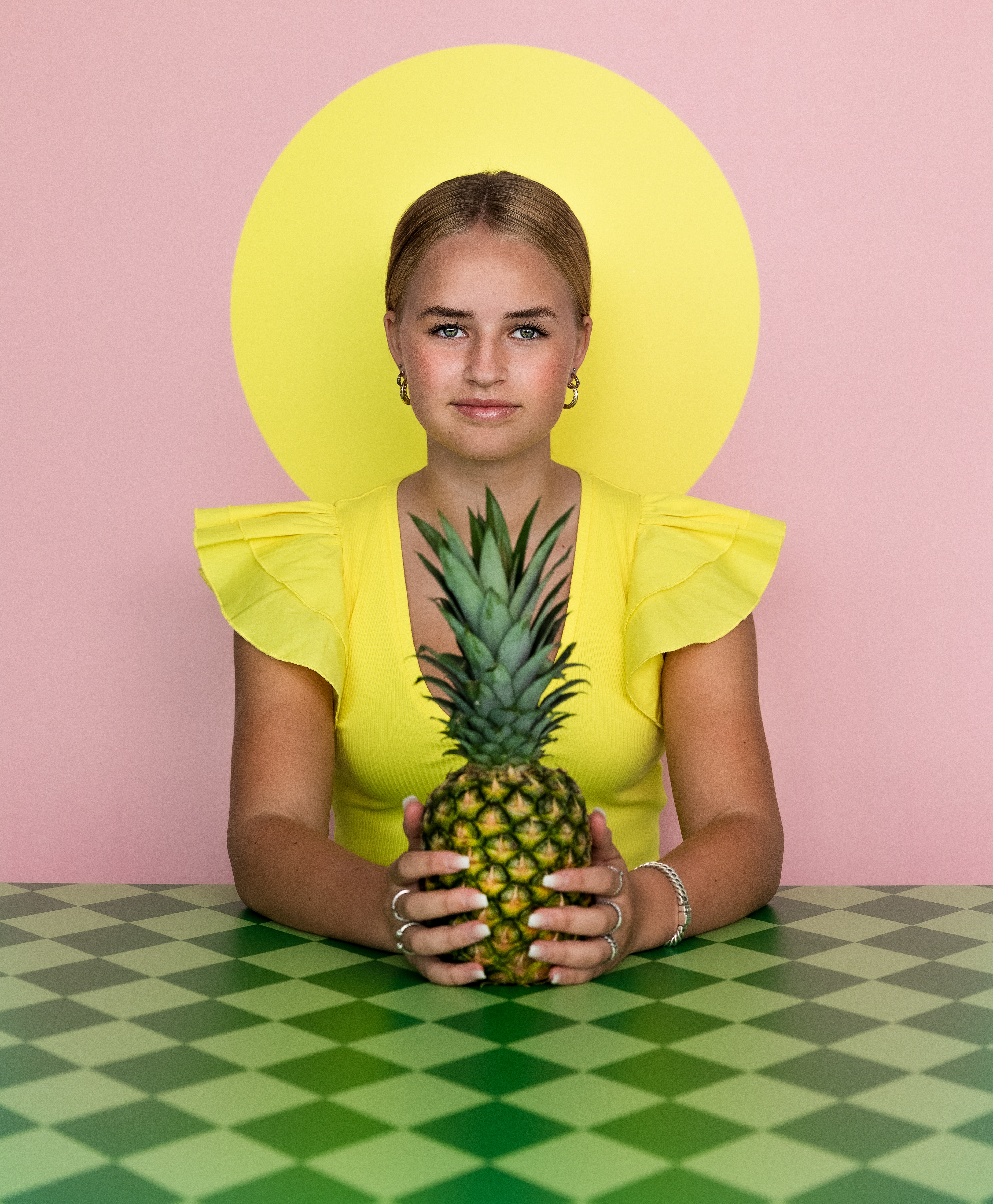 Stand tall, wear a crown and be sweet on the inside. Tiener meisje zit voor een roze achtergrond met gele stip. Deze stip zit precies achter haar hoofd en lijkt een aureool. Ze draagt in de zelfde gele kleur een top met korte mouwe.n. Ze zit aan een tafel met daarop een groene grafische print en ze houd een ananas vast die op de tafel staat. De foto is helemaal symmetrisch.
