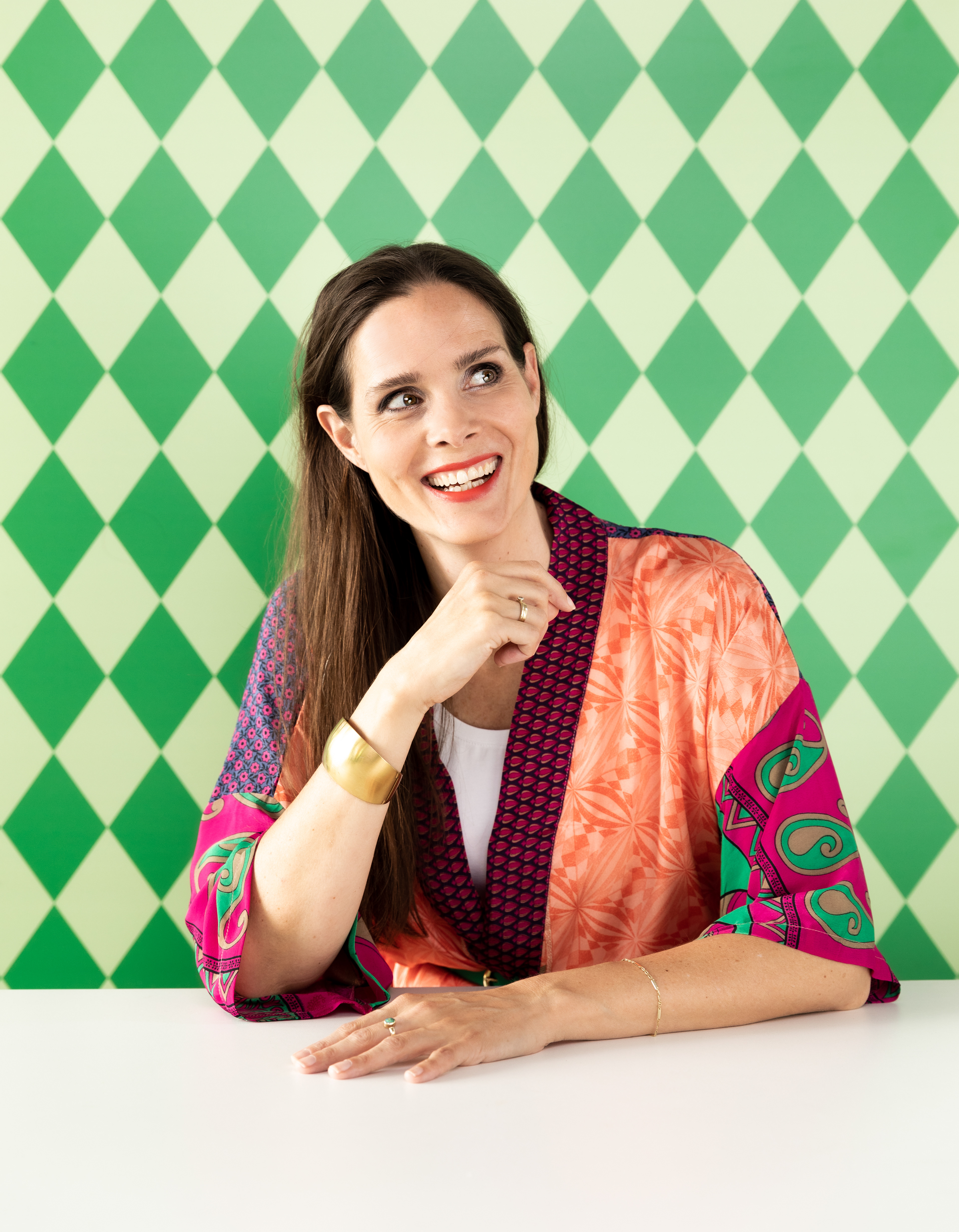 Portret van een lachende vrouw voor een grafische groene print. Ze draagt een kimno met verschillende kleuren en prints zittend aan een witte tafel