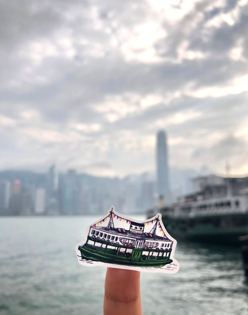 Hand holding sticker of a star ferry boat with background of Victorian harbour Hong Kong skyline with cloudy sky