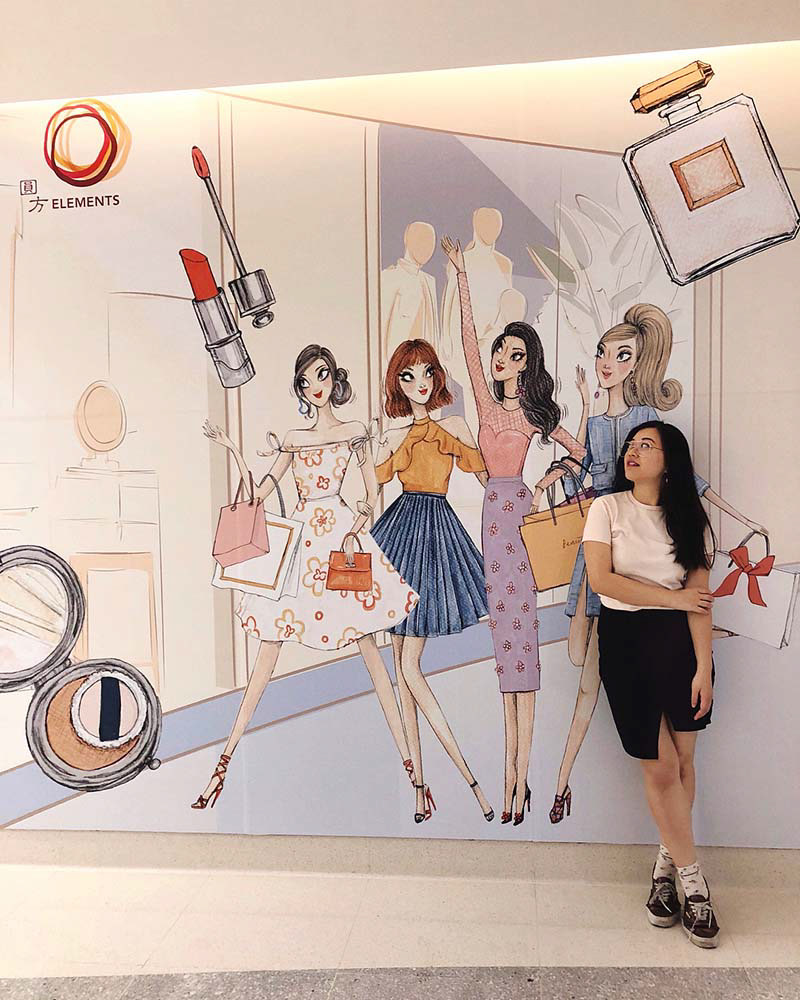 Chinese girl with long black hair in front of a mall display with an photo of fashion drawings