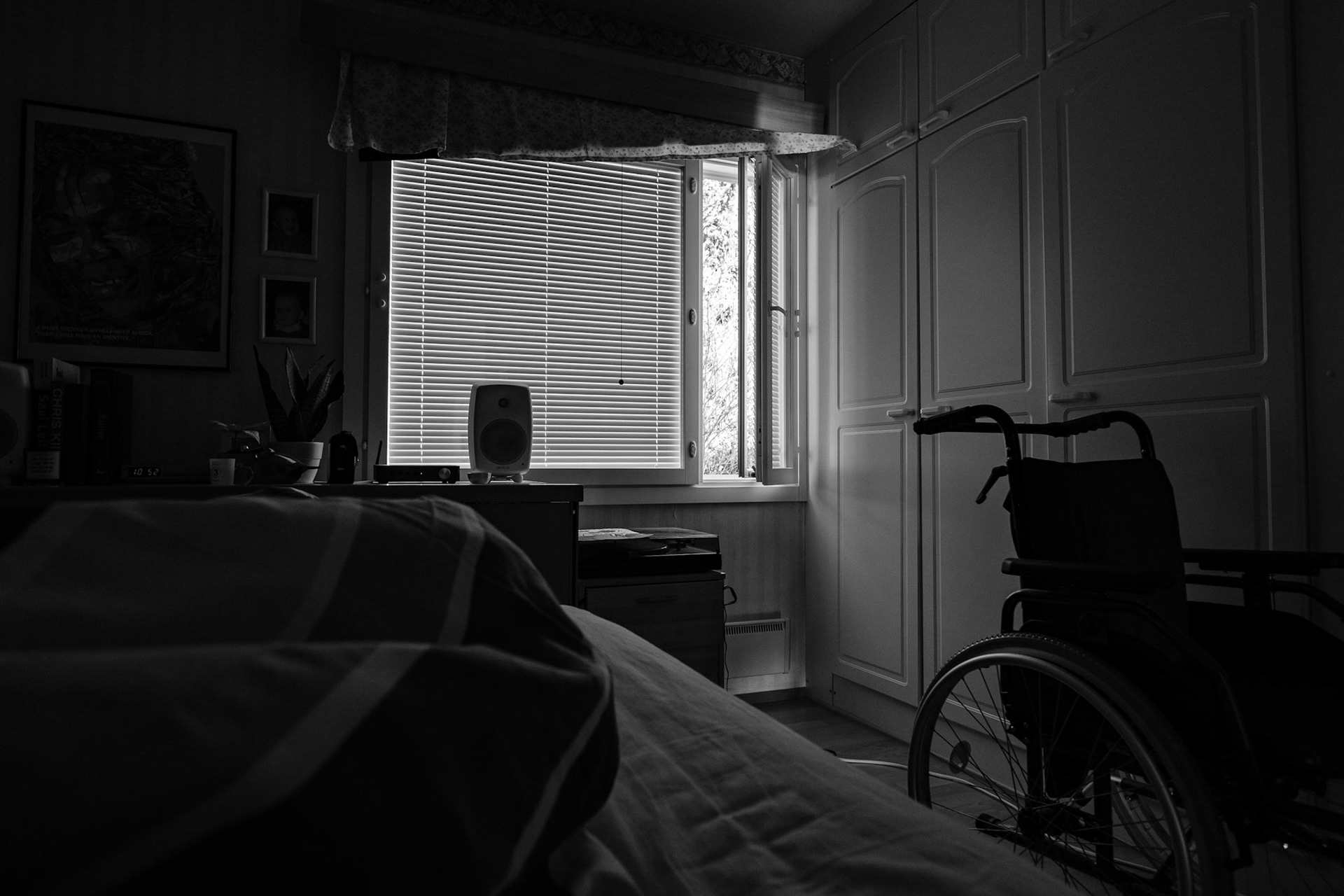 A black and white photo taken from Niko’s point of view while he lies in bed. The room is dark; the blinds are closed but the side window is open. A wheelchair is visible next to the bed.