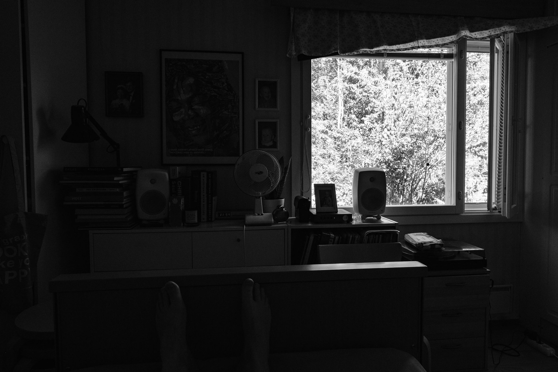 A black and white photograph of a view from a bedroom window to outside, with trees and vegetation filling the window view. The small side window is open. A sideboard has a fan on top of it.