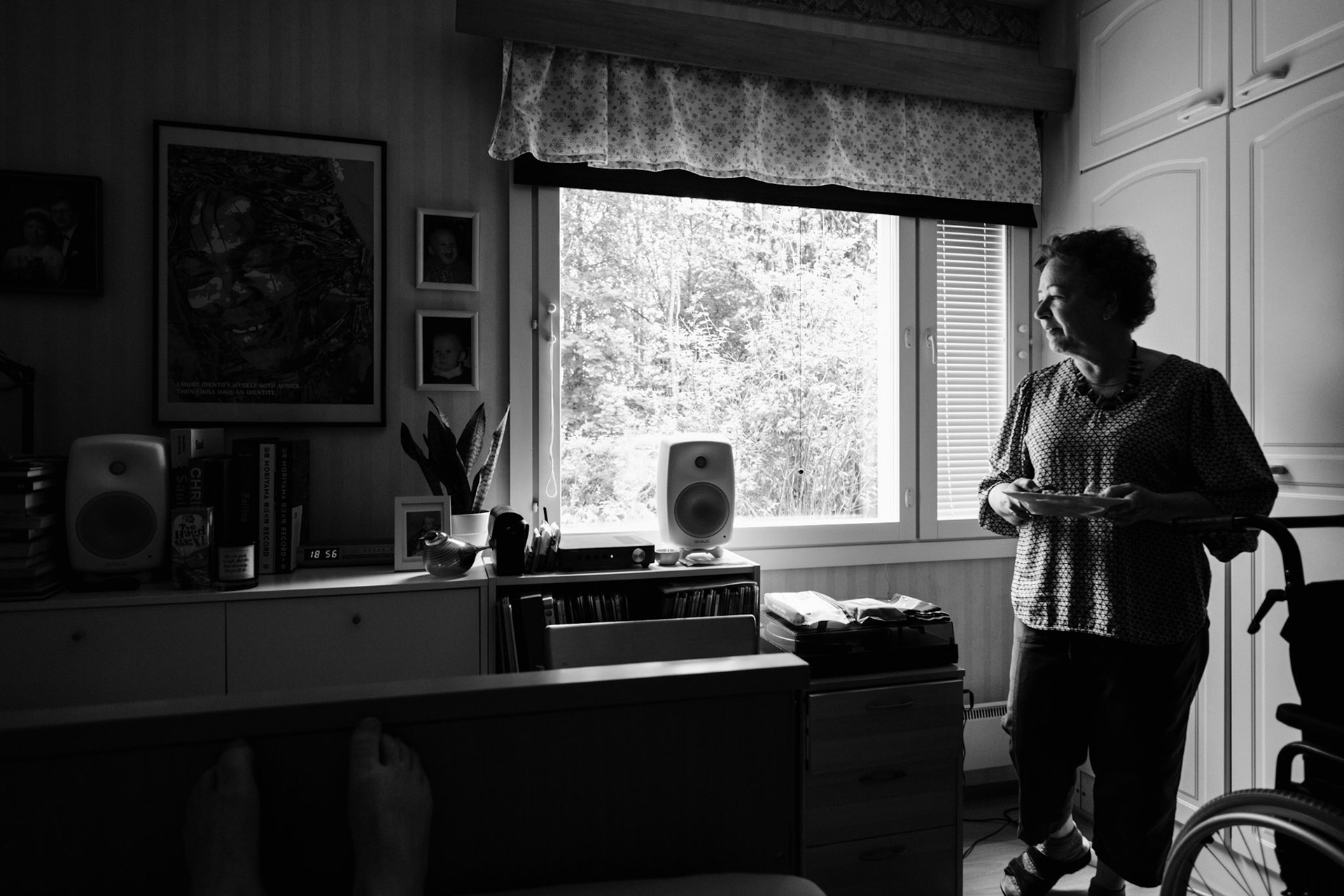 A black-and-white photo of a window view revealing a summer landscape. Inside the room, Niko’s mother has brought him his lunch. She is holding a plate in her hands. There is a turntable and a speaker, and various personal belongings on top of a sideboard.