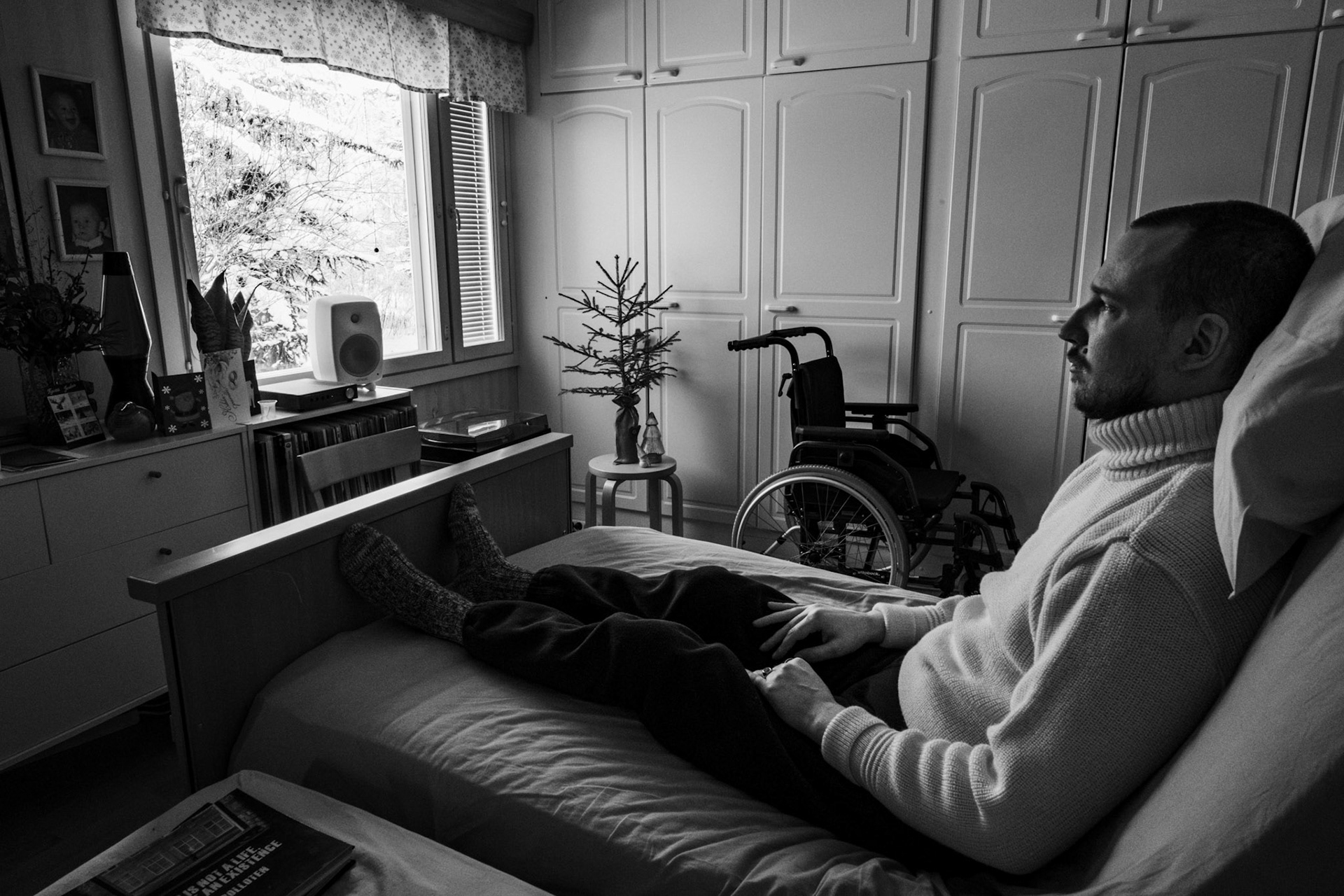 A black-and-white photo of Niko, a man in his 30s, sitting upright in a medical bed, looking outside a window. He’s wearing a thick woollen turtleneck sweater. The view from the window reveals a snowy landscape. In the background are a small Christmas tree on a stool and a wheelchair.