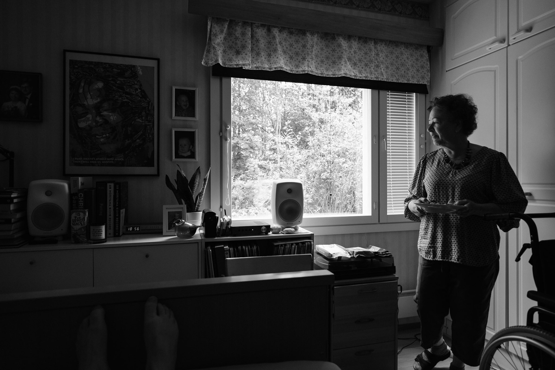 A black and white photo of a window view revealing a summer landscape. Inside the room, Niko’s mother has brought him his lunch. She is holding a plate in her hands. There is a turntable and a speaker, and various personal belongings on top of a sideboard.