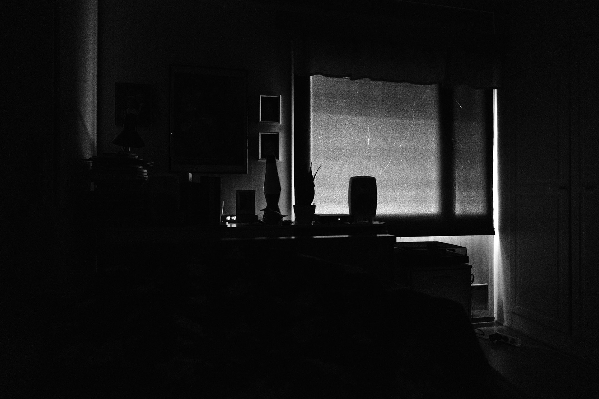 A high contrast black and white photograph of a completely dark bedroom. A long and narrow ray of light peaks through the corner of the curtain-covered window. The silhouette of the window is seen through the curtain.