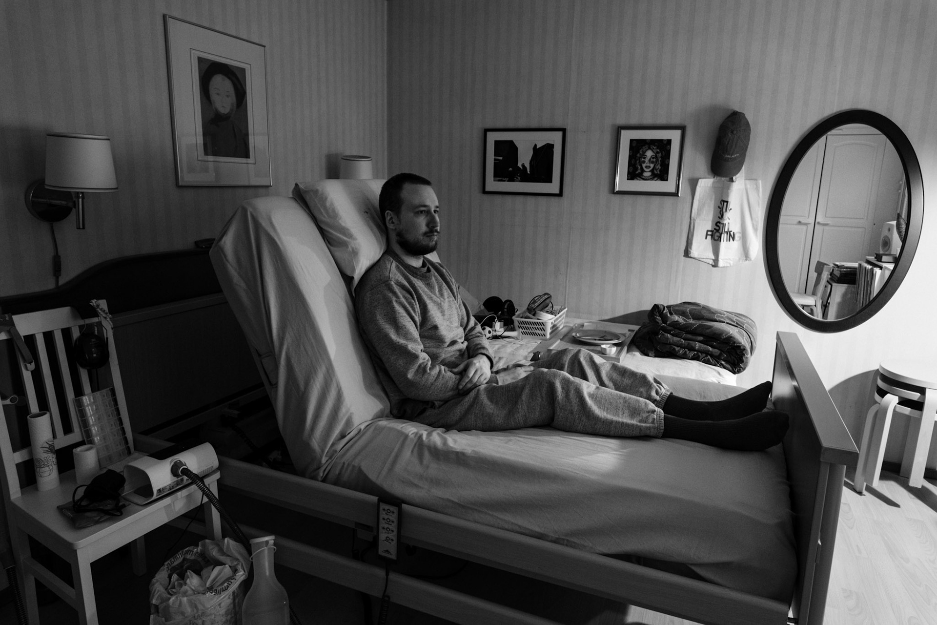 A black-and-white photo of Niko sitting upright in a medical bed, his arms crossed and he looks out of the window. The room has a dim light. A CPAP machine and medical equipment are beside the bed.