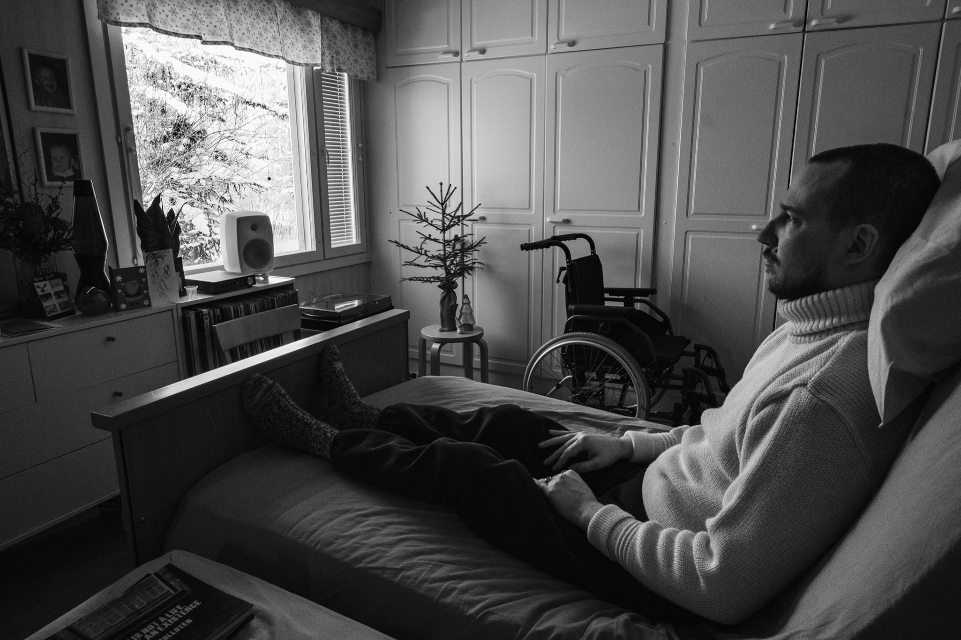 A black and white photo of Niko, a man in his 30s, sitting upright in a medical bed, looking outside a window. He’s wearing a thick woollen turtleneck sweater. The view from the window reveals a snowy landscape. In the background are a small Christmas tree on a stool and a wheelchair.